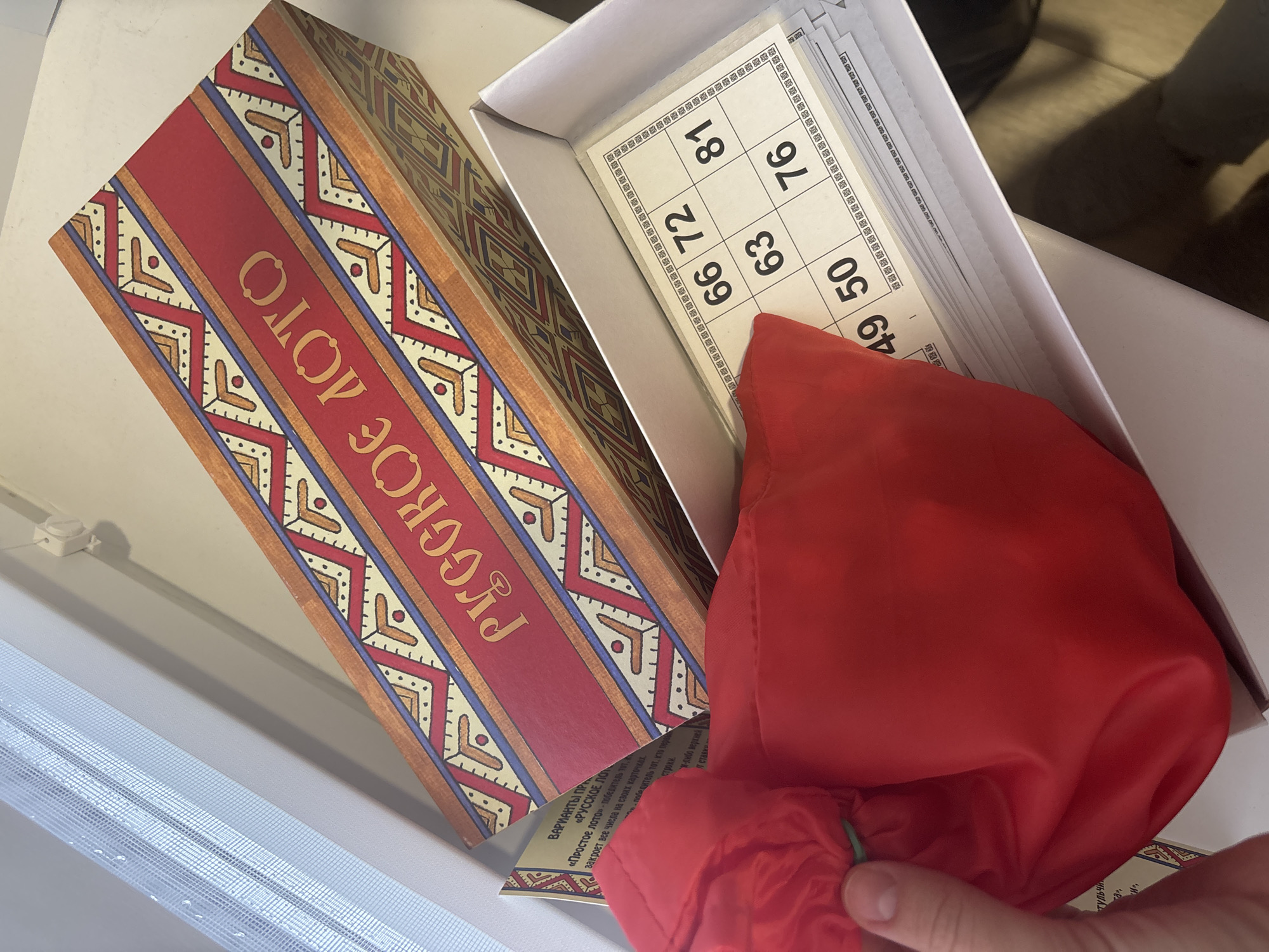 The image shows a "Russian Lotto" game set, likely in a home setting. The game box, with the name "Русское Лото" (Russian Lotto) printed on it, is next to an open box containing number cards. A red drawstring bag, presumably holding the number barrels, is partially out of the box. The game pieces and box are on a white surface that could be a windowsill or table. The image includes fragments of text near the bag. The OCR text confirms the name of the game "Русское Лото" and describes the rules. Given the name of the game, the reversed text on the box in Russian language, it is possible that the location is related to the Russian version of the game, perhaps the city of Chaikovsky, Russia.
