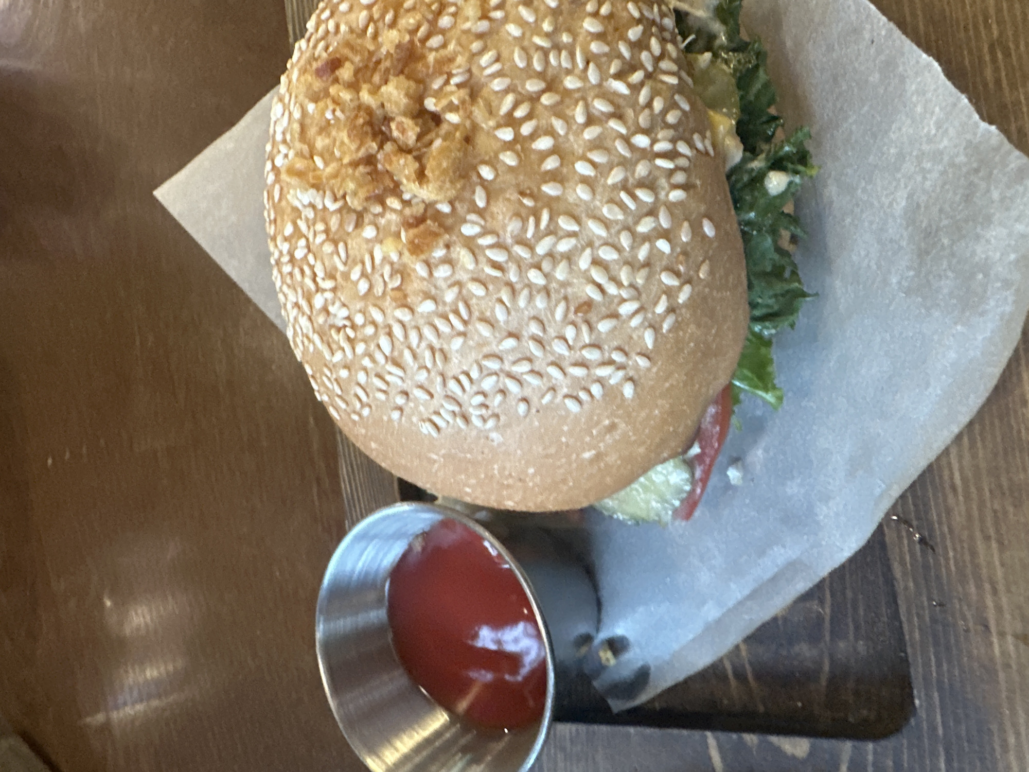 A close-up, overhead shot shows a burger and a small metal ramekin of ketchup on a wooden table. The burger is on a piece of parchment paper, which is on top of a dark wooden serving tray. The burger has a sesame seed bun, and some of the toppings are visible, including lettuce and tomato. The ketchup is a bright red color. The table surface is dark wood with a visible grain pattern. There are no people or discernible actions in the image. The lighting suggests an indoor setting, likely a restaurant. Blagoveshchensk, Russia is not visually represented in the image.