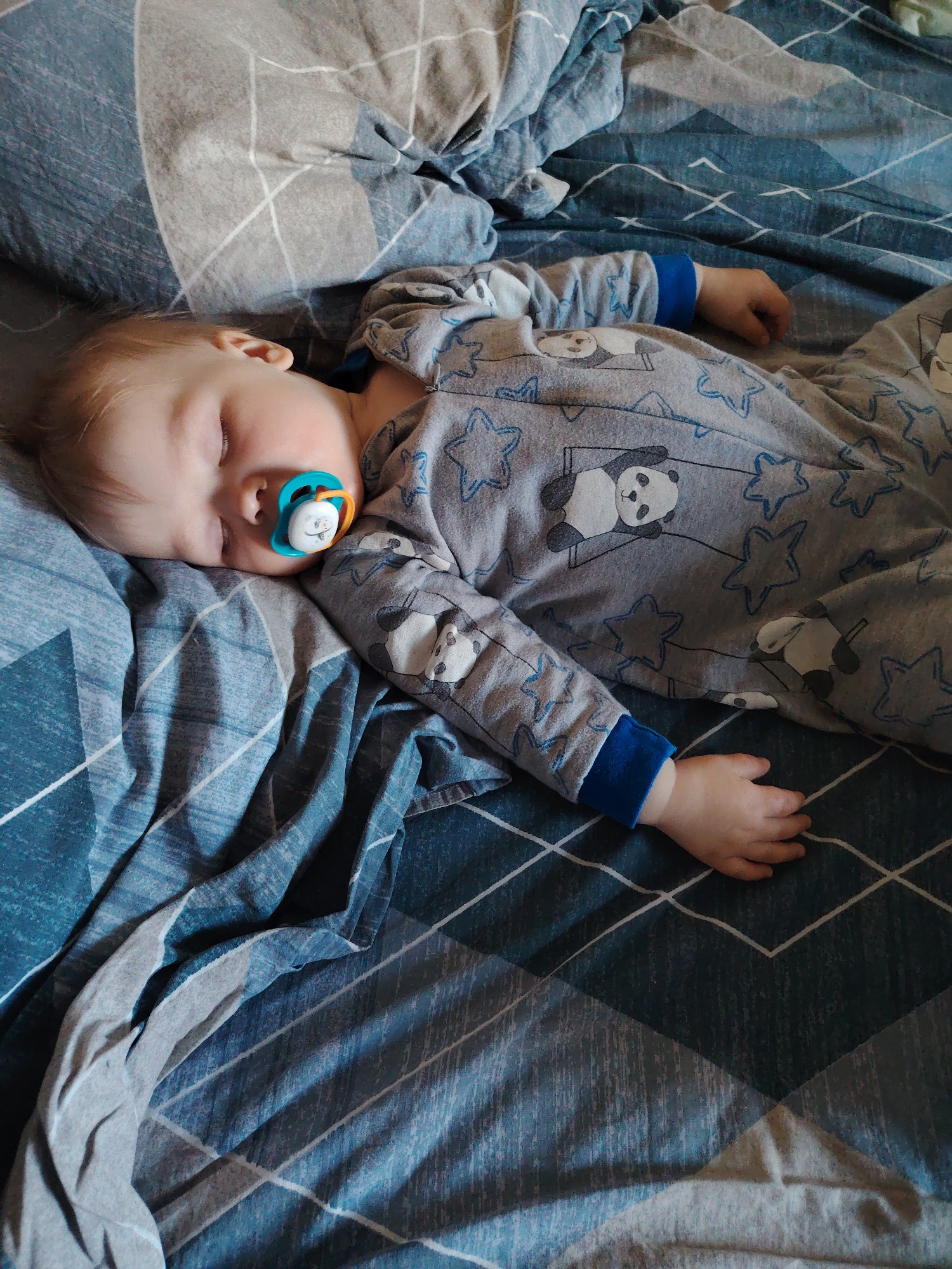An overhead shot captures a baby or toddler peacefully asleep on a bed. The child, with light blonde hair and fair skin, lies on its right side with eyes closed, and a blue, orange, and white pacifier in its mouth. The pacifier features a small, light-colored animal design on its white central part.
The baby is dressed in grey pajamas patterned with blue stars and cartoon pandas. The sleeves have distinct blue cuffs. Its arms are bent, with one hand resting near its chest and the other outstretched on the bedding.
The bedding consists of a duvet cover and possibly a sheet, both exhibiting a geometric pattern in shades of blue and grey, featuring squares, diamonds, and linear designs. The textures appear soft and comfortable.
The lighting in the image is soft and even, suggesting an indoor setting during the daytime or in a well-lit room, contributing to a serene and calm atmosphere. There is no visible text on any of the items in the image. Given the indoor setting, no specific visual cues within the image indicate the specified location of Chaikovsky, Russia.