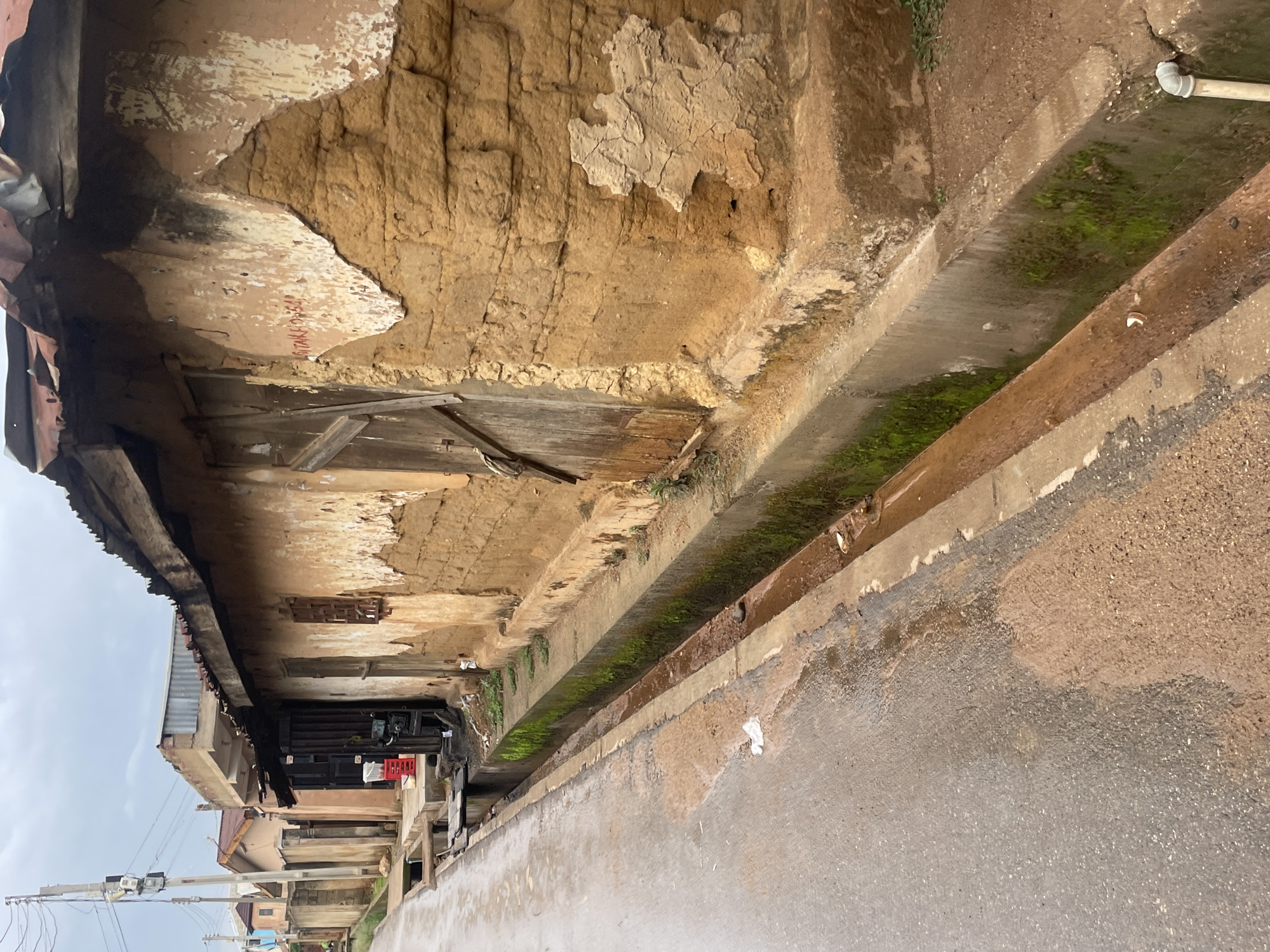 The image depicts a street scene in Jos, Nigeria, characterized by a worn, earthy-toned building constructed from mud bricks with sections of plaster peeling off. The building has a corrugated metal roof, some of which appears damaged or rusted. Adjacent to the building is a concrete gutter running alongside a paved road. The gutter contains some water and green moss, suggesting recent rain or a constant flow of water. The road itself appears wet, with patches of exposed earth or mud. In the background, other buildings and a utility pole with wires are visible. The overall impression is one of a developing urban environment with signs of age and potential disrepair.