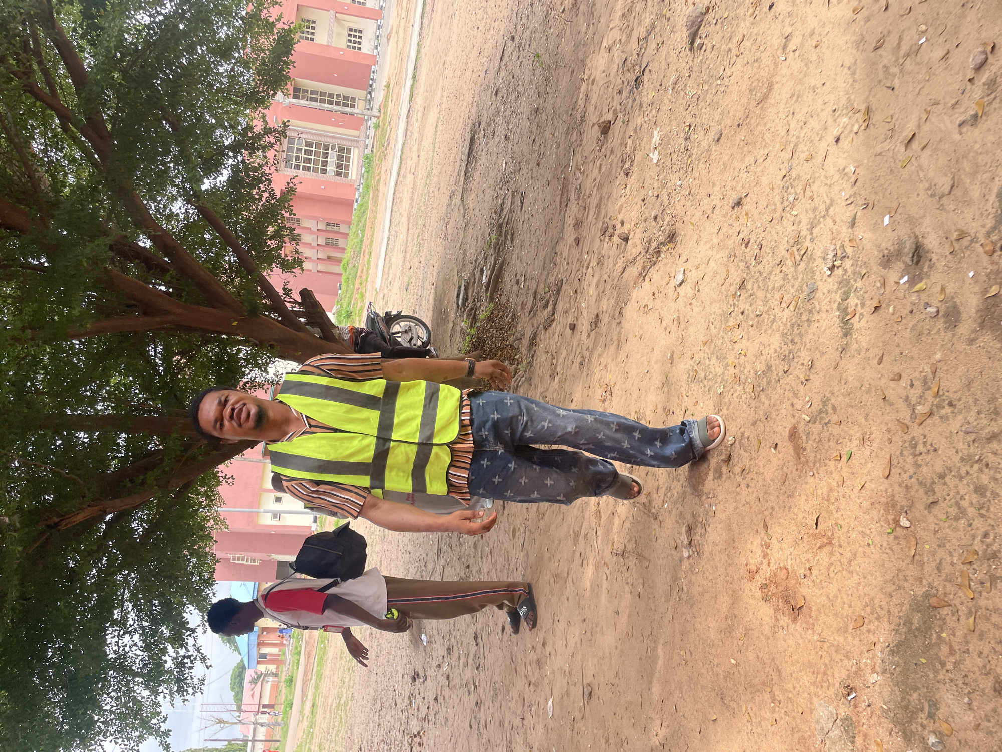 The image depicts an outdoor scene on a dirt path in Lokoja, Nigeria, under the canopy of a large tree. Two young men are the primary subjects. In the foreground, a man wearing a bright yellow safety vest over a striped shirt and patterned blue jeans walks towards the camera. He is looking directly at the camera with a slight smile. To his left and slightly behind him, another young man is walking away from the camera, wearing a white t-shirt with a red trim, brown trousers with a red stripe, and black sandals. He has a black backpack slung over his right shoulder. In the background, a light pink building with numerous windows and a darker pink trim is visible. A portion of a motorcycle is parked to the left of the foreground subject, partially obscured by the tree. The ground is covered in dry, reddish-brown dirt, scattered with fallen leaves and small stones. The overall impression is that of a casual encounter on a campus or open area within the city.