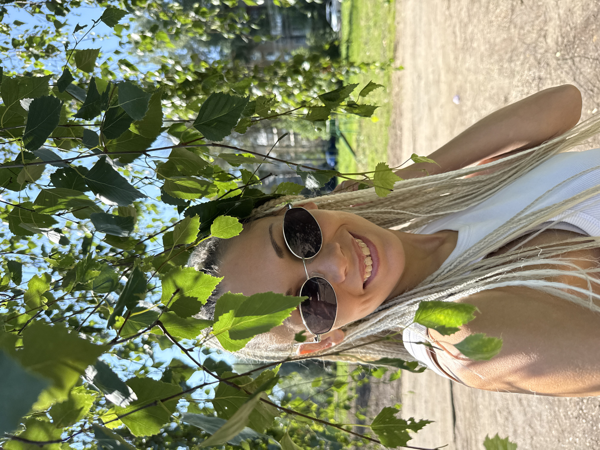 A medium close-up shot captures a smiling woman with blonde braids and sunglasses outdoors, with birch tree branches and leaves in the foreground and a blurred background of a park with buildings and cars. The woman is wearing a white top and is positioned slightly to the right of the frame. Sunlight filters through the leaves, creating dappled light on her face and shoulder. The ground appears to be dirt or gravel. The location is identified as Rusanovo, Russia.