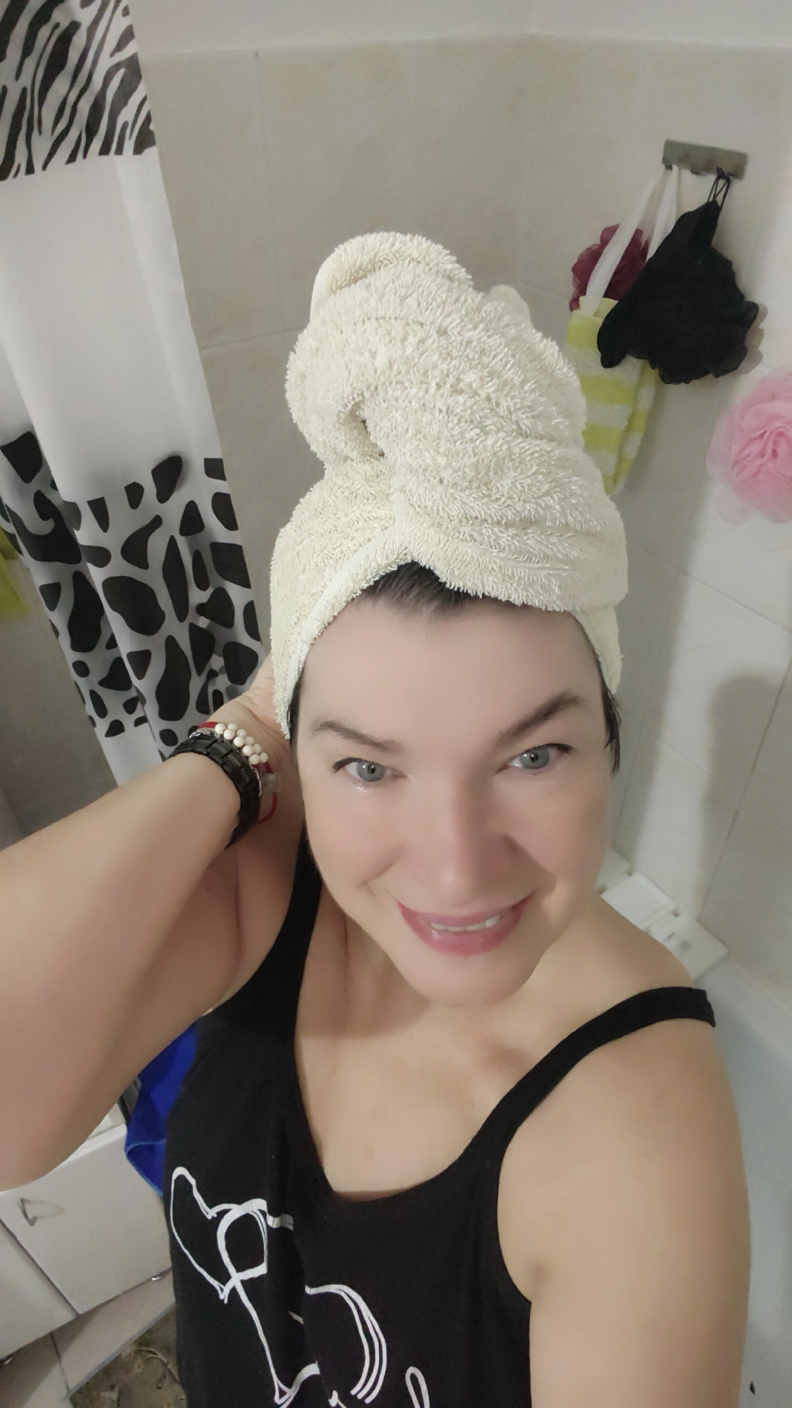 A woman is in a bathroom in an unknown city. She is wearing a black tank top and has a white towel wrapped on her head. She is smiling and has her hand up near her head, possibly adjusting the towel. Behind her is a shower curtain with a black and white patterned print. On the wall there are some bath accessories hanging. The scene appears to be a selfie taken in a residential bathroom.