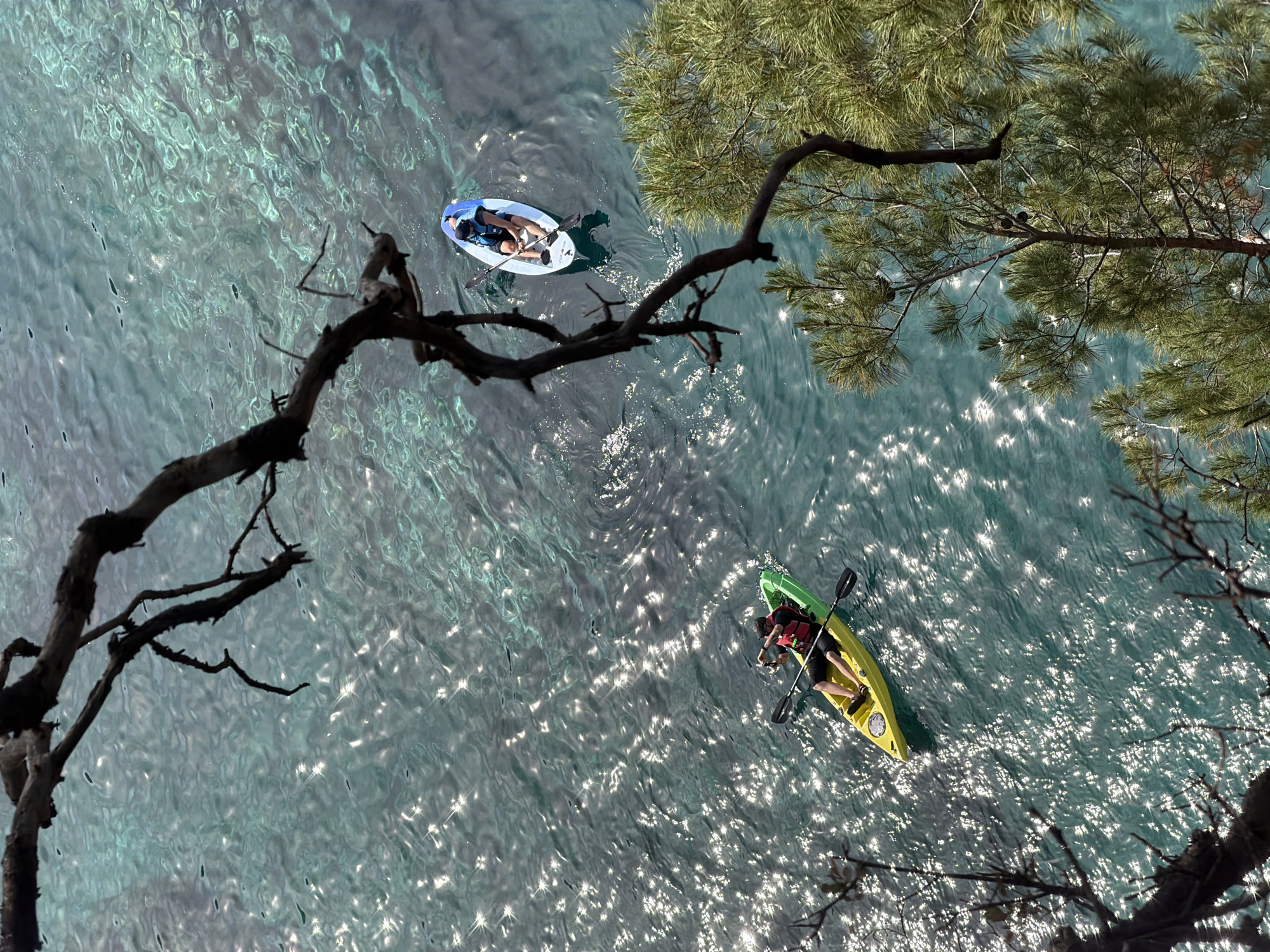 Aerial view of Petrovac centar, Montenegro, showing two people on watercraft. One person lies prone on a light blue paddleboard. The second person is paddling a yellow and green kayak and wears a red life vest. The water is turquoise and reflects sunlight. Tree branches frame the top and left side of the image.