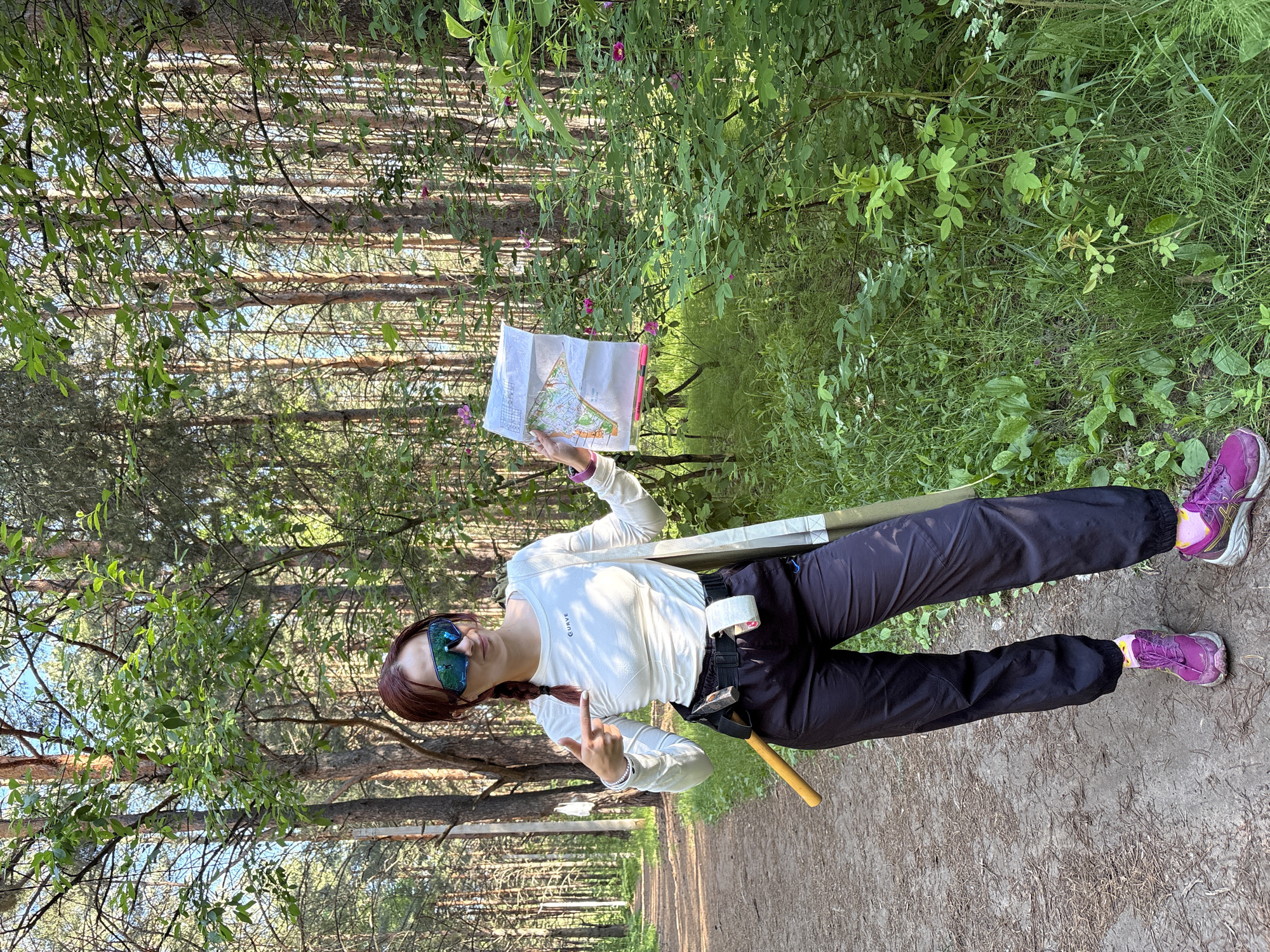 A young woman with auburn hair styled in a braid stands on a dirt path in a forest. She is wearing a white long-sleeved shirt with the word "CURVE" printed on it, dark pants, bright purple athletic shoes with yellow accents, and blue-tinted sports goggles. Around her waist, she has a black belt with an axe attached to her left hip. She holds a folded map in her right hand and points to it with her left index finger. The forest appears to be composed of pine trees and dense green foliage. The path is lined with grass and fallen pine needles. The scene suggests an outdoor activity, possibly orienteering or hiking, within a natural environment. The provided location of Chaikovsky, Russia, indicates the geographical setting of this image.