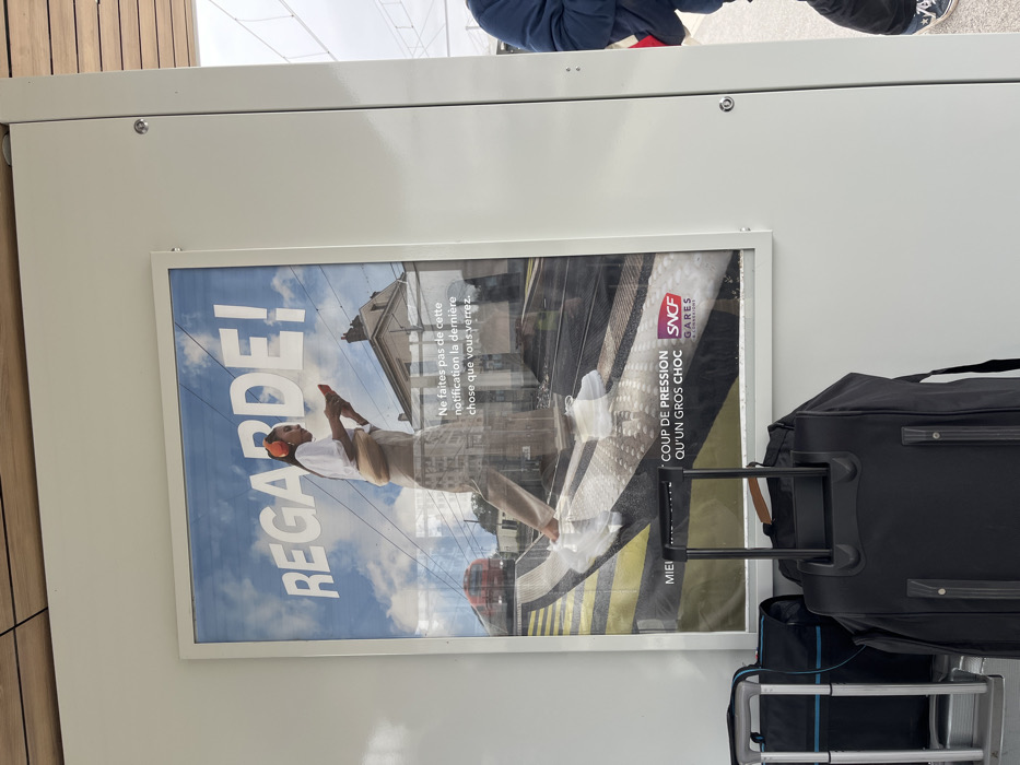 The image depicts a framed railway safety advertisement poster mounted on a clean, white vertical panel at a train station in Dijon, France. The poster features a young woman with dark hair wearing orange over-ear headphones, a white short-sleeved top, light-colored pants, and white sneakers, looking down at a red smartphone in her hands. She is depicted walking on a train platform with tactile paving, with a blurred red train approaching from the left and a building in the background, all under a blue sky. The large white text at the top of the poster reads "REGARDE!" (Look!). Below the main image, text states: "Ne faites pas de cette notification la dernière chose que vous verrez." (Do not let this notification be the last thing you see.) Further down, "COUP DE PRESSION QU'UN GROS CHOC" (A push/pressure rather than a big shock/impact) is visible next to the "SNCF Gares & Connexions" logo. In the immediate foreground, two pieces of luggage are visible: a black soft-sided suitcase with an extended black telescopic handle in the center, and the silver handle and part of a black and blue bag to its left. On the far right, the lower right torso, arm, and leg of an individual wearing a dark blue jacket and black and white sneakers are partially visible. The scene is brightly lit.