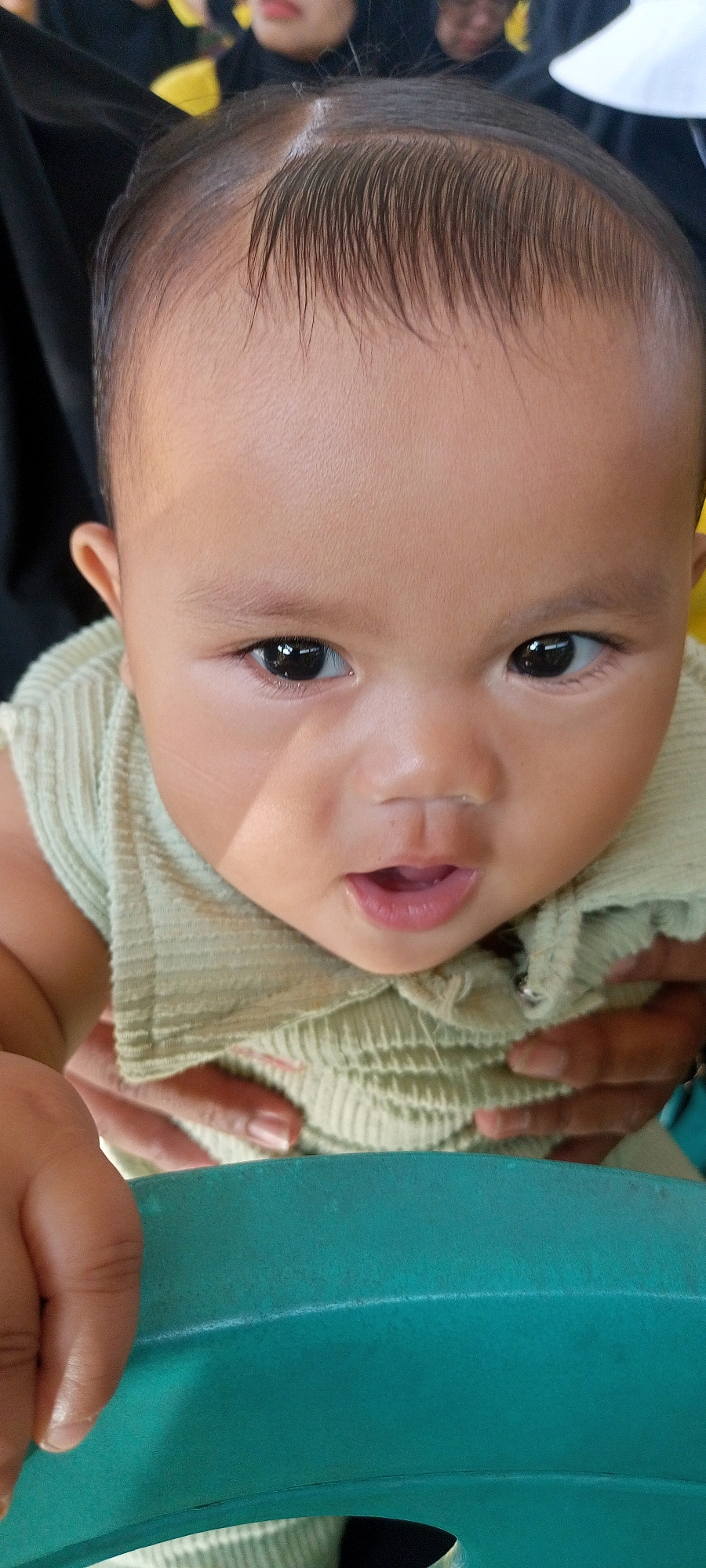 The image shows a close-up, head-on view of a baby's face. The baby has dark hair, large dark eyes, and an open mouth. The baby is wearing a light green, ribbed shirt. Two hands are visible, one on each side of the baby's torso, holding the baby. The background is blurred and dark, with hints of yellow and white. A teal-colored object, possibly a chair or table edge, is in the foreground. The image appears to be taken indoors, with the baby being held by someone.