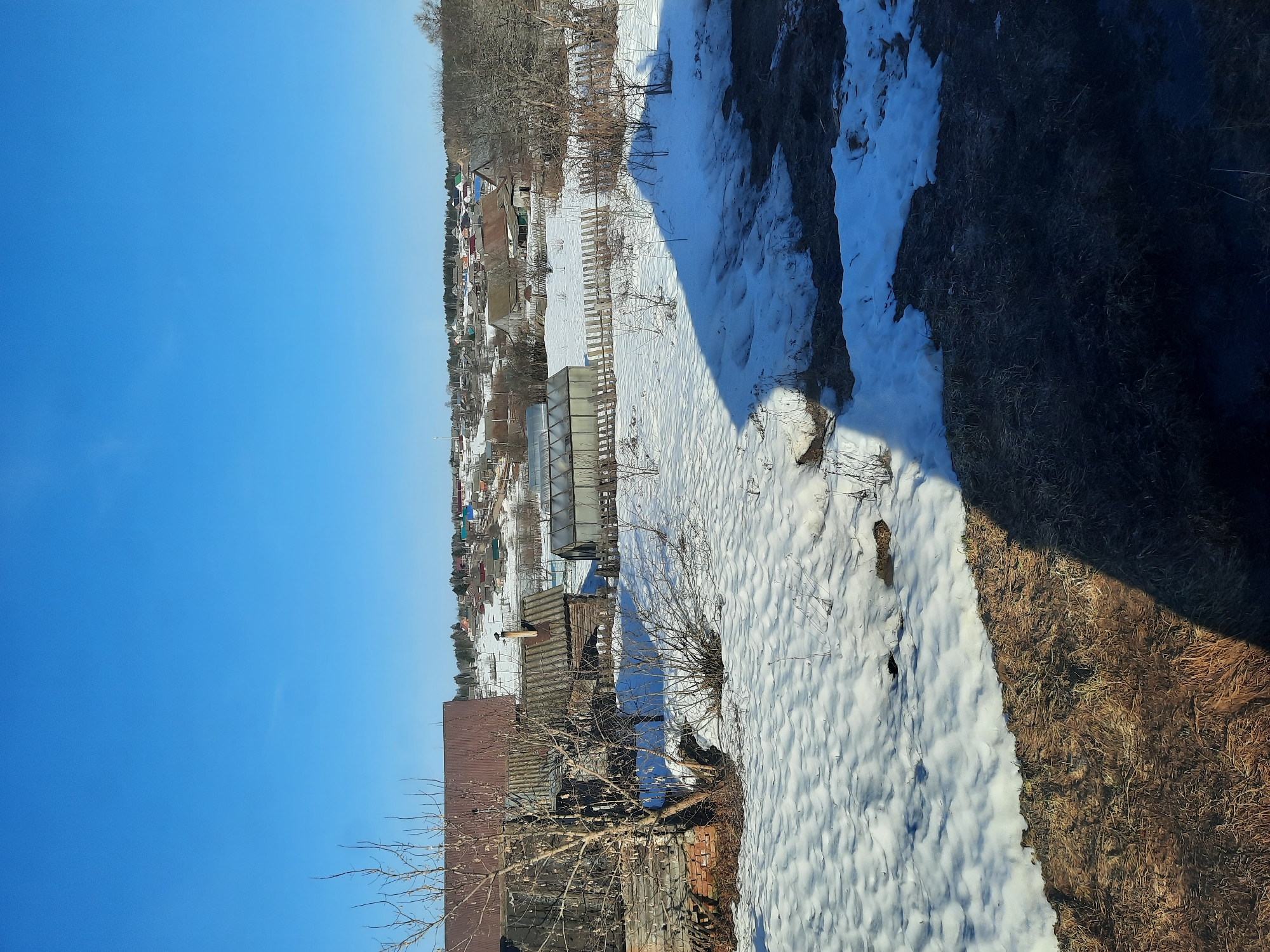 The image shows a winter scene in what appears to be a rural or suburban area of Krasnogorskoye, Russia. Patches of snow cover the ground, particularly in the areas between buildings and near vegetation. Several houses with pitched roofs are visible, some with brick or wood exteriors. A greenhouse-like structure is also present, suggesting agricultural activity. Bare trees and bushes dot the landscape. To the right, a shadow and a steep, grassy embankment with some snow are visible, perhaps indicating the image was taken from an elevated position or building. The sky is clear and blue, suggesting a sunny day. Overall, the scene portrays a typical Russian village landscape transitioning from winter to spring.