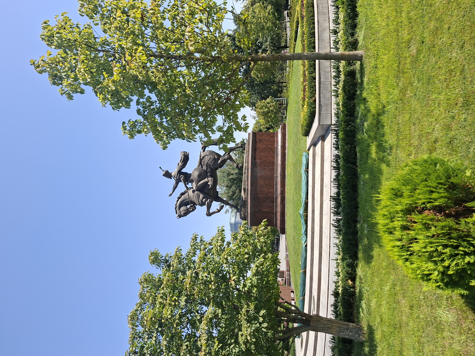 A bronze equestrian statue of Amir Timur stands on a granite pedestal in a public park in Tashkent, Uzbekistan. The statue depicts Amir Timur on horseback, raising his right arm in a gesture of leadership, with a sword in his left hand. The base of the statue is surrounded by lush green grass and manicured bushes. Two trees flank the statue, with their leaves fully bloomed. The sky is clear and blue. In the background, modern buildings can be seen peeking through the trees, indicating an urban setting. The overall scene conveys a sense of history and pride in Uzbekistan's heritage.