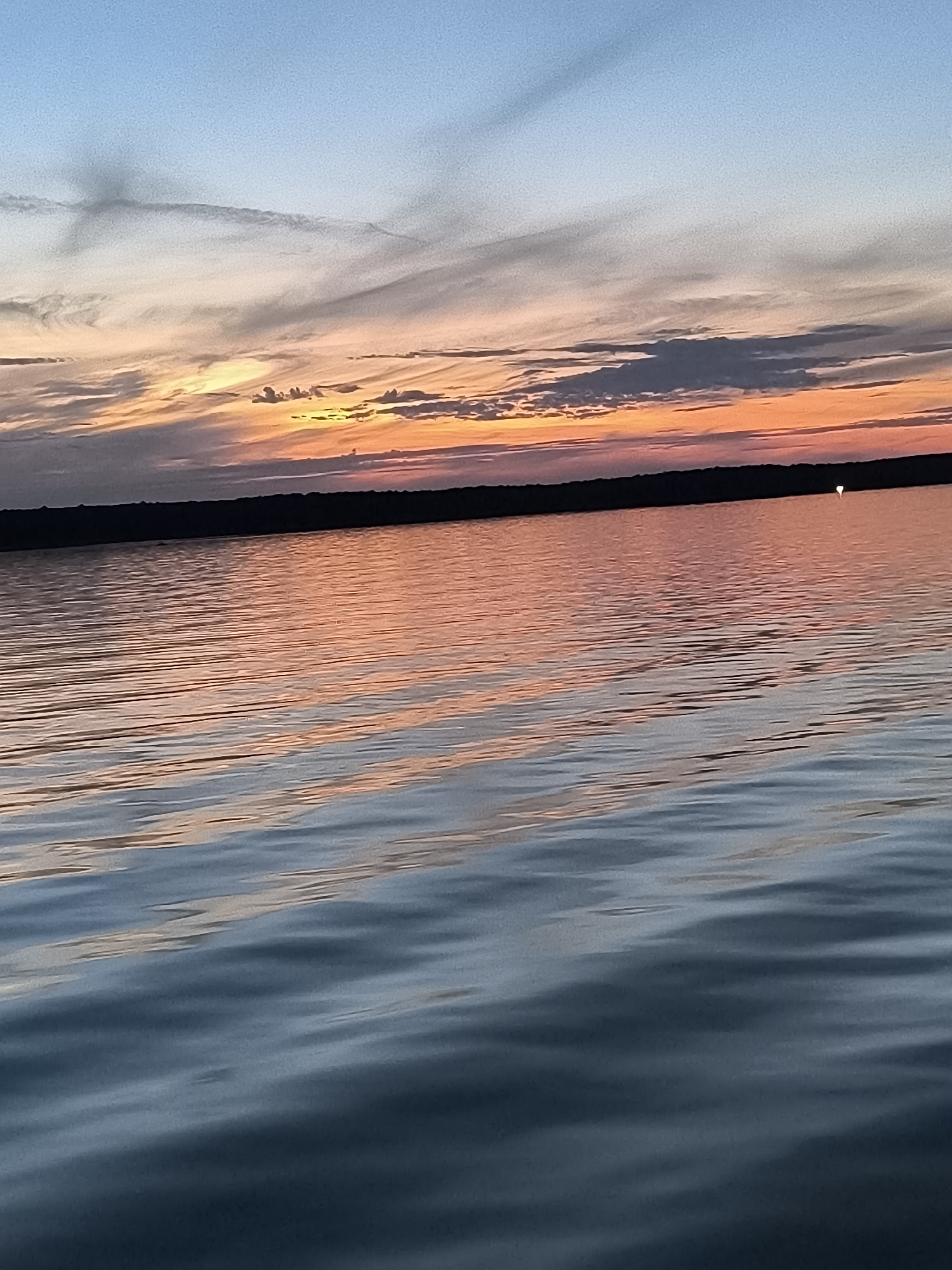 The image depicts a serene natural landscape at sunset, likely over a body of water, with a dark, tree-lined horizon. The sky transitions from a clear blue at the top to vibrant hues of orange, pink, and grey clouds closer to the horizon, indicating the setting sun. The water reflects these colors, with streaks of orange and pink shimmering across its surface, which is gently rippled. In the far distance, on the horizon line, a small, bright white light is visible, possibly a distant boat or a navigational marker. The overall atmosphere is peaceful and tranquil. Given the description of Neringa, Lithuania, this scene could be situated over the Curonian Lagoon or the Baltic Sea, where such sunset vistas are common. The image does not contain any people, specific objects of interest, or actions occurring.