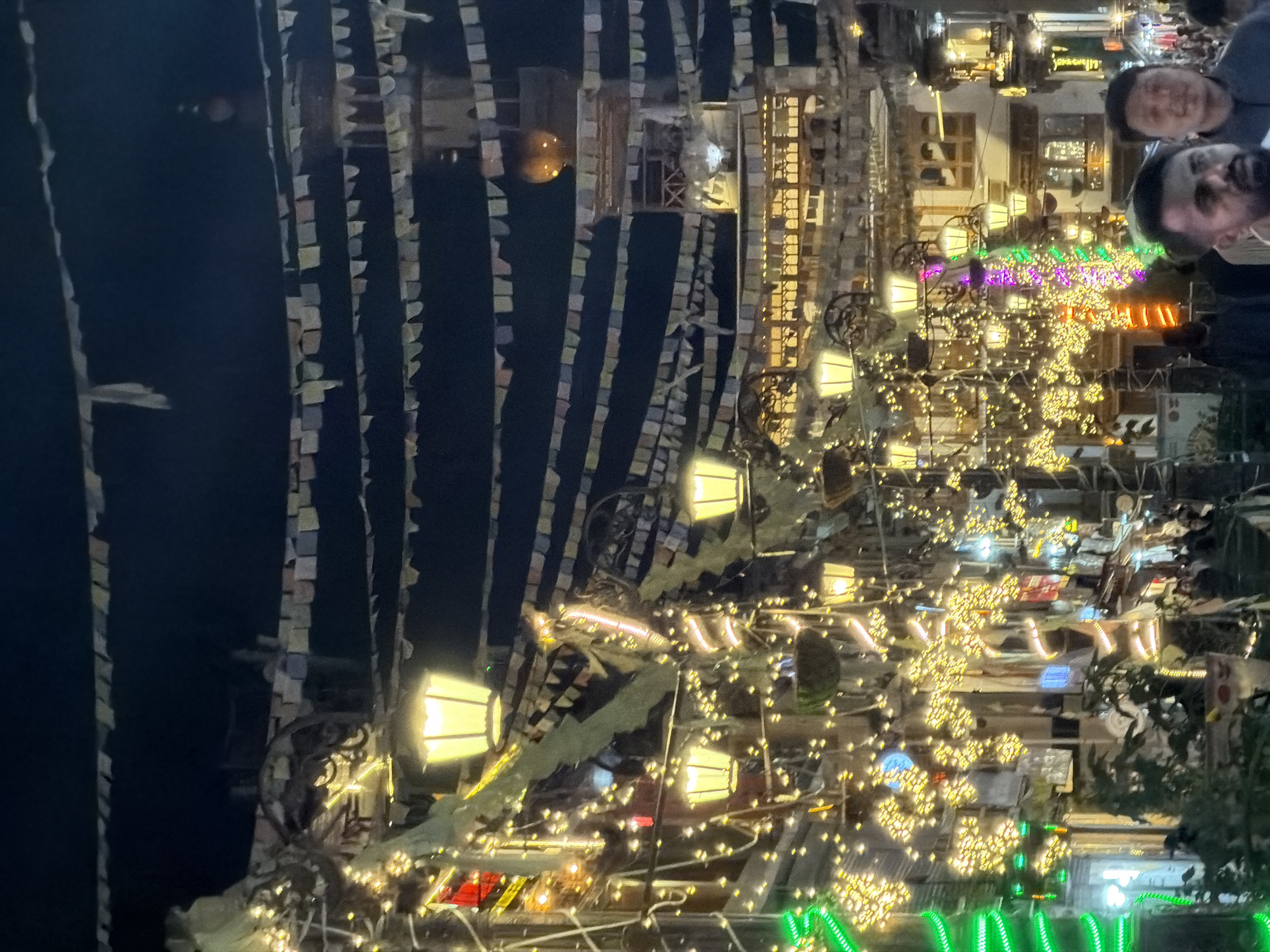 The image depicts a street scene at night in Leh, India, illuminated by numerous decorative lights. The street is lined with buildings adorned with strings of yellow, white, and green lights, creating a festive atmosphere. Prayer flags are strung across the street, a common sight in Himalayan regions. In the background, a building with architectural elements resembling a stupa or minaret is visible. Several people are walking along the street, some appearing to be tourists. The overall impression is one of a lively and culturally rich urban environment at night.