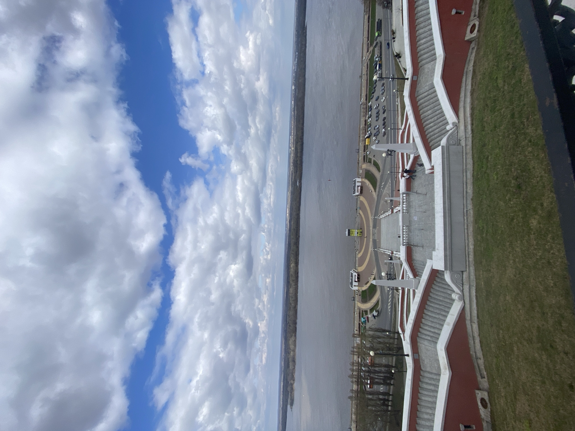 This image, viewed from an elevated perspective, captures a scenic view of a wide river, a grand urban embankment, and a partly cloudy sky in Nizhny Novgorod, Russia.
In the foreground and to the right, a prominent architectural feature dominates: a multi-tiered staircase system, likely part of the Chkalov Staircase, descends towards the river. The stairs are characterized by their white balustrades, grey steps, and sections of red walling, flanked by grassy slopes. A dark metal railing is visible on the extreme right, suggesting the photograph was taken from a balcony or viewing platform.
Below the staircase, a paved embankment road stretches along the riverbank. Numerous cars are parked along the road, and some traffic is visible. Streetlights and sparsely leaved trees line the thoroughfare. Further along, a patterned circular plaza or turnaround area is present, with small white structures that might be kiosks or information points. A few individuals can be seen near the base of the stairs, with two figures appearing to be seated on a bench or railing.
The expansive river occupies the midground, its surface appearing calm. A distant, treelined landmass forms the opposite bank. A small, dark object, possibly a boat or buoy, is visible on the water's surface.
The upper portion of the image reveals a vibrant blue sky dotted with scattered, fluffy white cumulus clouds, indicating fair weather during the daytime, likely in early spring or late autumn given the appearance of the trees. Visible text on signs and structures along the embankment is too small or blurry to be deciphered.