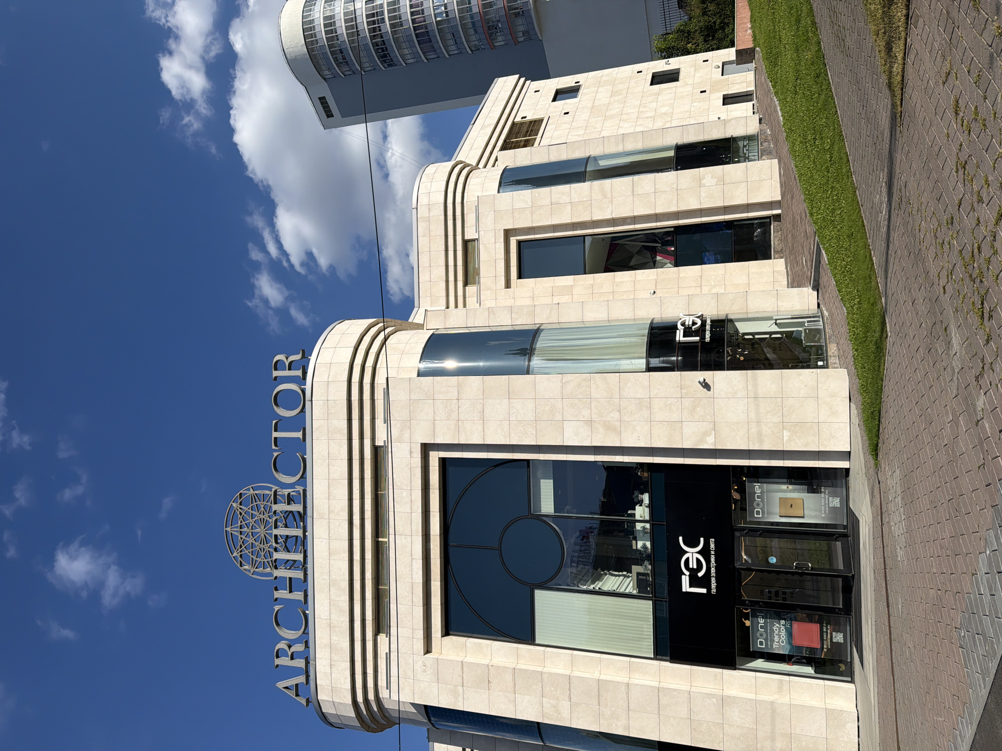 The image shows the facade of the "ARCHITECTO" building, a modern commercial structure in Yekaterinburg, Russia. The building is constructed with beige stone tiles and features large, dark-tinted windows. The name "ARCHITECTO" is displayed prominently in large, metallic letters above the main entrance, accompanied by a stylized circular logo. Below the name, a black sign reads "ГЭС" (GES) in white lettering, with smaller text underneath that appears to be "галерея электрики и света" (gallery of electricity and light).
On the ground floor, there are storefronts with glass doors and displays showcasing products. One display features a red item and text indicating "Trendy Colors RGB". Another display shows a gold item and text with a phone number and website "donel.su". The building is situated on a slight incline, with a paved walkway in the foreground and a grassy slope to the right. In the background, a tall, modern building with a curved facade and many windows is visible, as well as a clear blue sky with scattered white clouds. The overall scene is bright and appears to be taken during daylight.