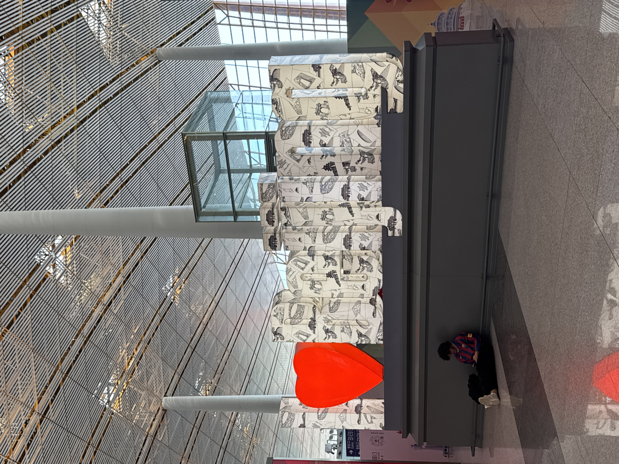 The image depicts an interior space, likely an airport or large public building in Chaoyang District, China. The architecture features a high, vaulted ceiling with a complex grid-like structure. Below this, a stack of luggage with distinctive patterned covers is situated against a gray partition. An oversized red heart-shaped object is placed near the base of the luggage stack. A person wearing a striped jersey and dark pants is sitting on the floor near the partition. The floor is a light-colored tile or stone. Natural light enters the space through the architectural grid above.