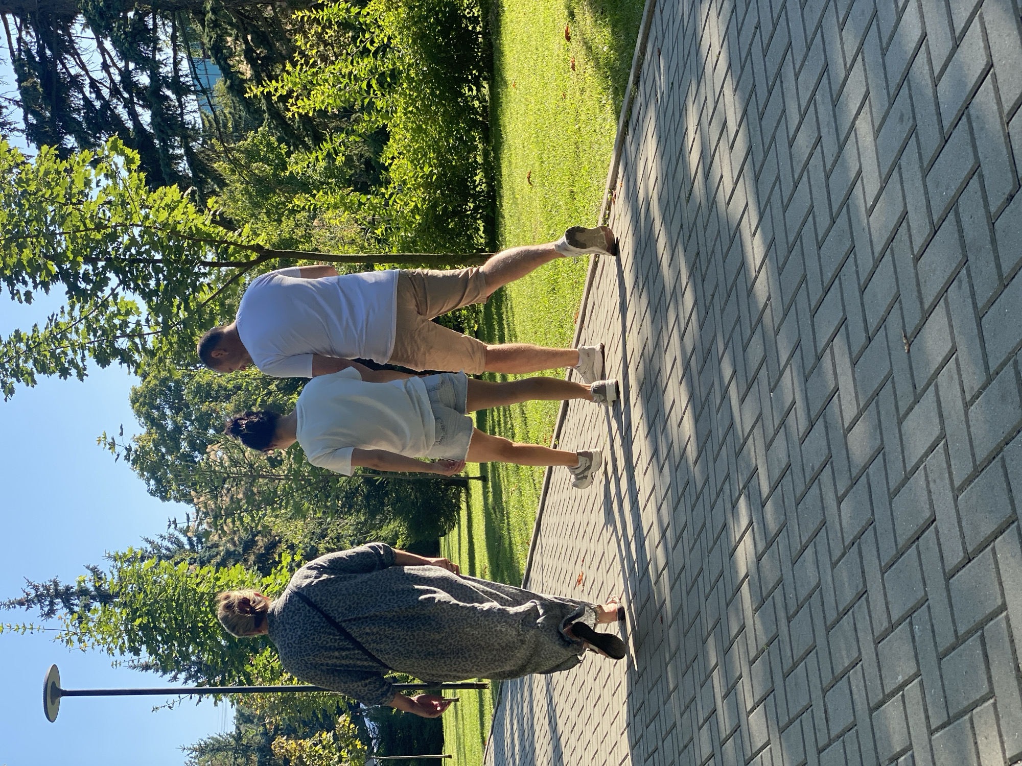 In an outdoor setting, three individuals walk along a paved pathway. The person on the left, a woman with her hair pulled back, wears a long, patterned gray dress and sandals. To her right walks a younger woman with her hair in a bun, dressed in a white t-shirt and denim shorts. On the far right, a man in a white t-shirt and light-colored shorts walks with a slight limp. The pathway is constructed of gray rectangular bricks laid in a herringbone pattern. Beyond the pathway, a well-maintained green lawn is visible, dotted with fallen leaves. Lush green trees and bushes form the background, with a clear blue sky above. The lighting suggests it is a sunny day, casting distinct shadows from the foliage onto the pathway. The overall scene depicts a casual walk in a park-like environment. The specific location in Chișinău, Moldova, cannot be determined from the provided image.