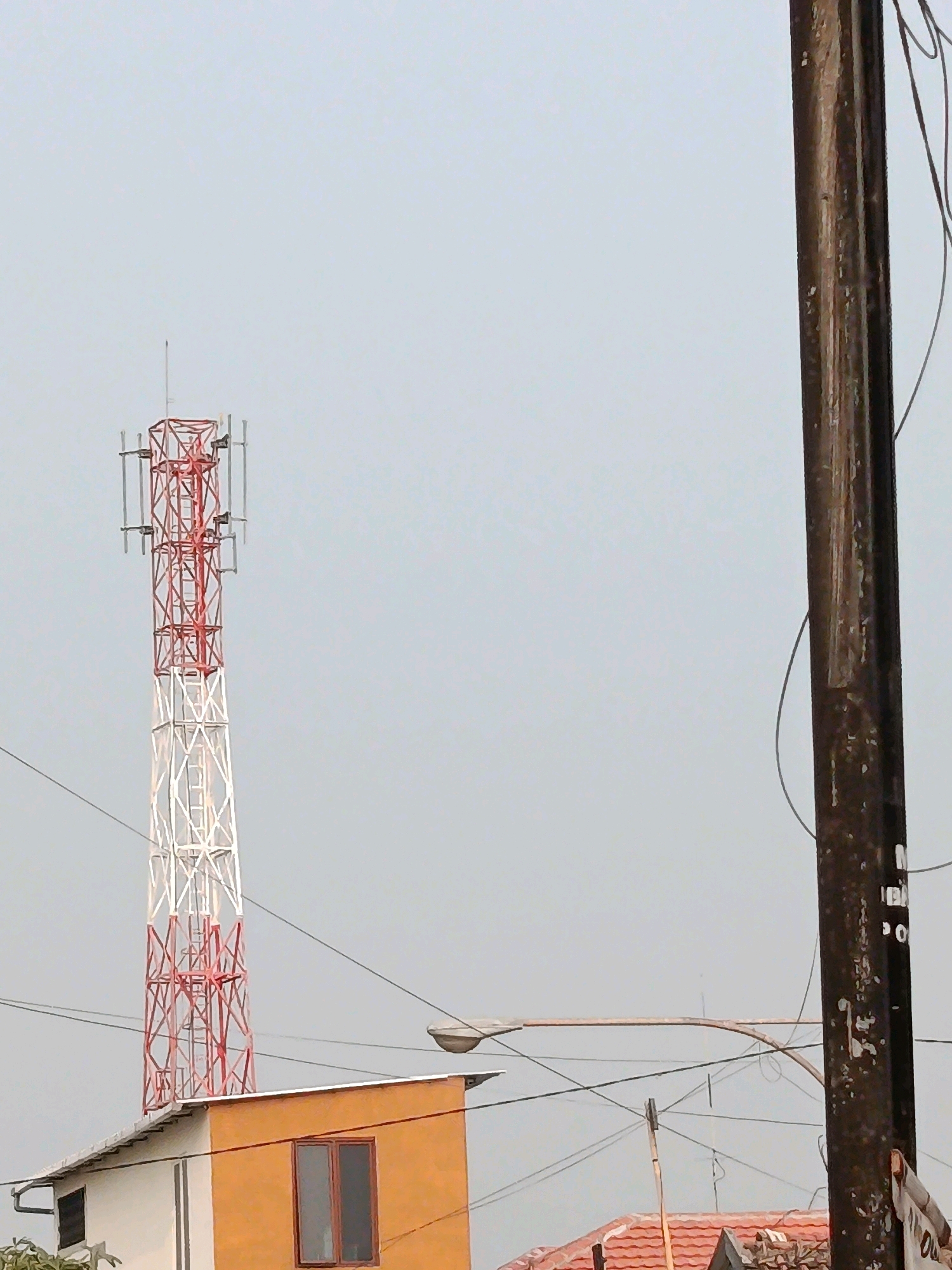 The image depicts an outdoor scene in the city of Jobokuto, Indonesia, dominated by a tall, red and white lattice telecommunications tower situated to the left of a dark, weathered utility pole on the right. The tower is equipped with antennas. Below the tower, a building with an orange and white facade is visible, featuring a dark-paned window. A street light extends from a pole towards the right side of the frame, passing over the building. Power lines crisscross the sky, connecting various structures. The sky is overcast and a light gray color. The foreground shows the roof of a building with red tiles.