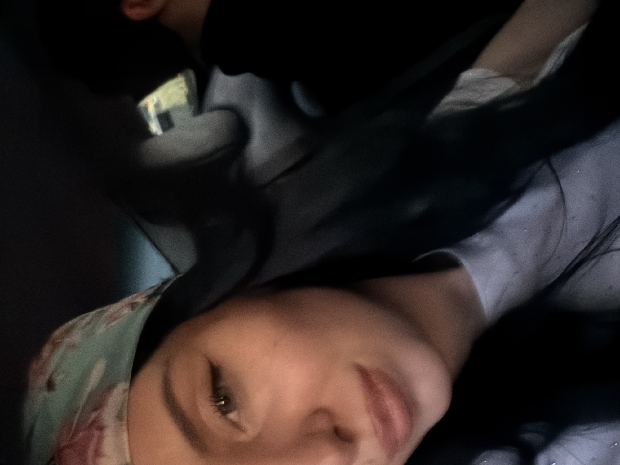 The image captures a close-up of a young woman with dark hair, wearing a headscarf with a floral pattern. She is positioned in the foreground, with her left eye and mouth visible. Her makeup includes winged eyeliner. She appears to be in the passenger seat of a vehicle, with the dark interior of a car visible behind her, including a headrest and a glimpse of another person in the background. The lighting suggests it might be daytime, possibly with some shade. The setting is not explicitly identifiable as Tbilisi, Georgia, as there are no specific landmarks or contextual clues within the image to confirm this location.