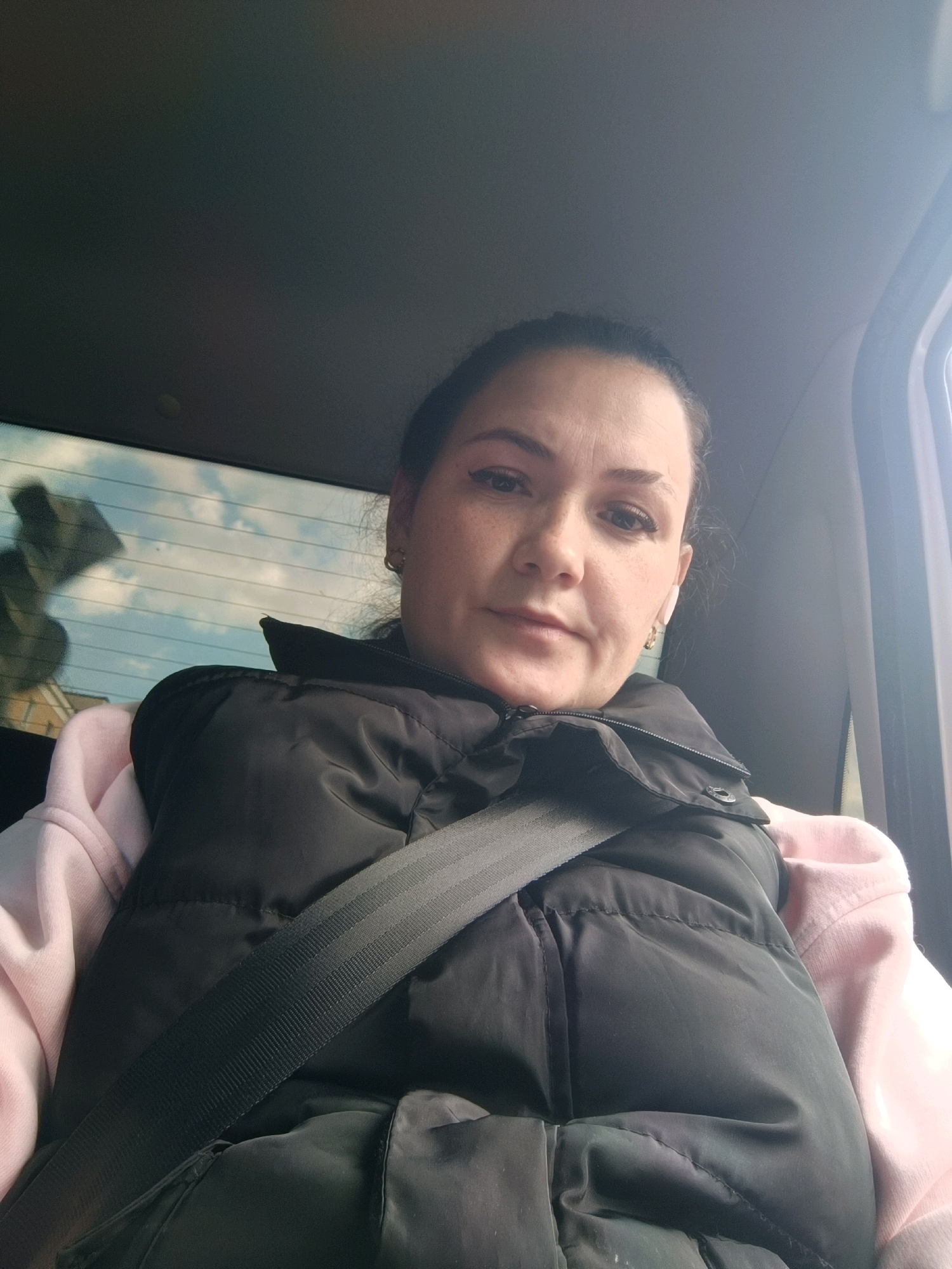 A young woman is sitting in the back seat of a car, wearing a pink hoodie and a black puffer vest. She is looking at the camera and appears to be taking a selfie. The seatbelt is visible across her chest. Through the rear window, a cloudy sky and buildings can be seen. The interior of the car is dark, with light entering from the side window. The image was taken in the city of Perm, Russia.