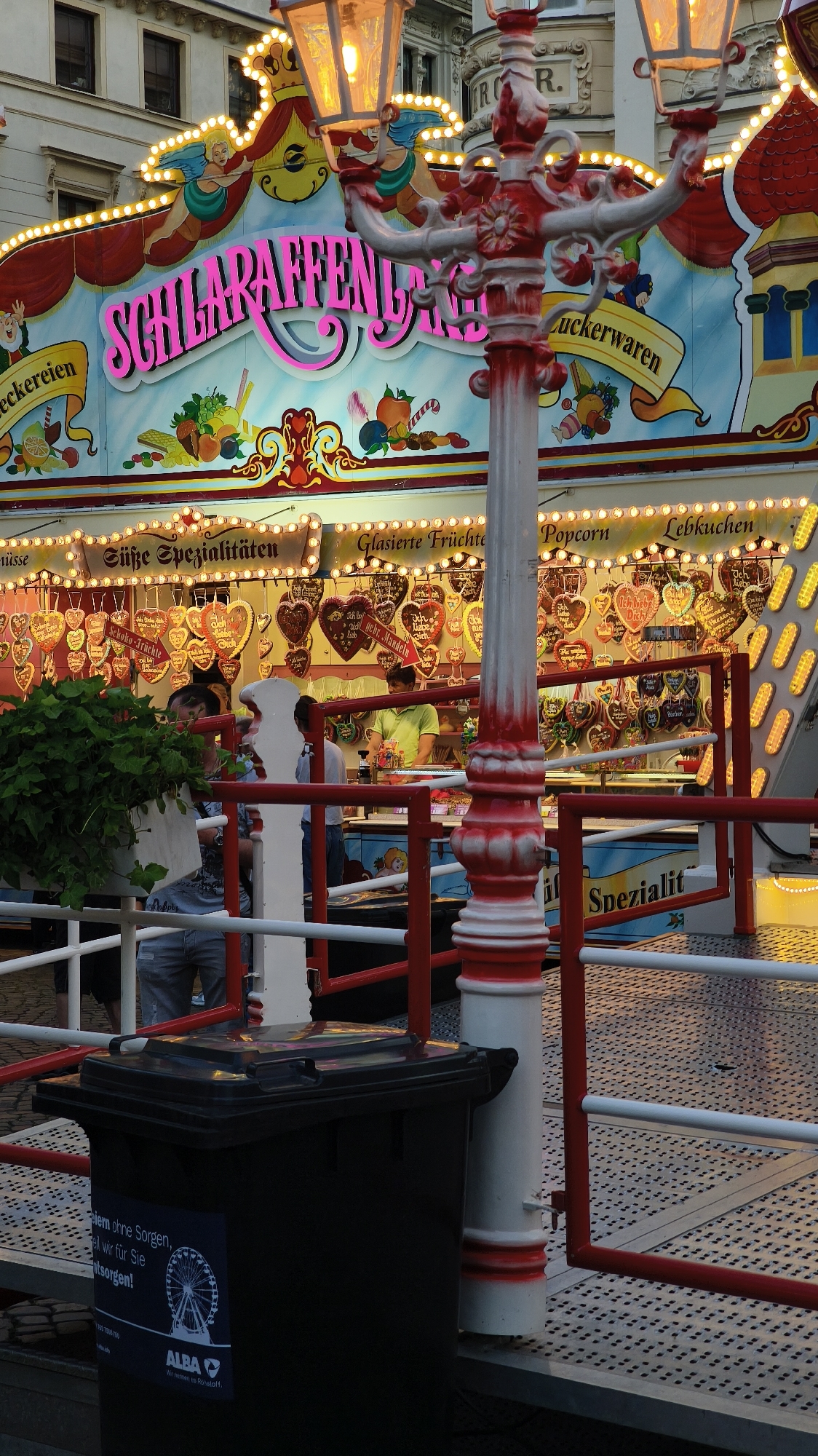An outdoor scene depicts a festive stall at a carnival or fair. The stall's facade is brightly decorated with a banner that reads "SCHLARAFENLAND" in large pink letters, flanked by images of cherubs and cartoon characters. Below this, a display showcases an assortment of goods, including heart-shaped cookies and various candies. In the foreground, a black trash bin with a sticker featuring a Ferris wheel and "ALBA" logo is visible. A red and white striped pole, part of the fair's decorative lighting, stands to the right of the bin. Several people are present, including a vendor in a green shirt behind the counter and patrons standing near the stall. The overall environment suggests a lively, public gathering.