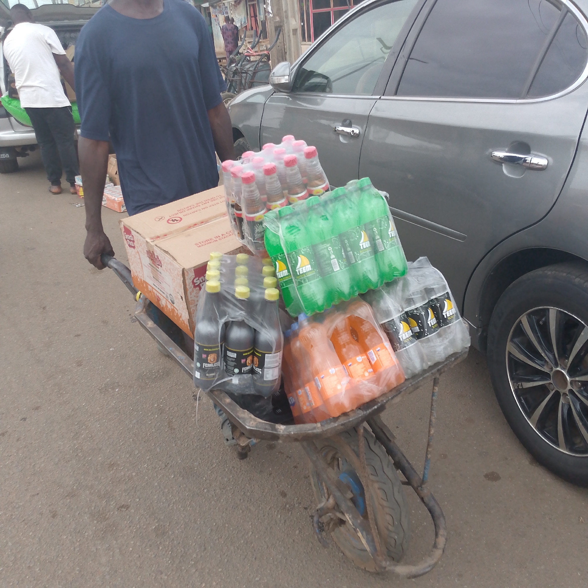 A man in Bukuru, Nigeria, is pushing a wheelbarrow filled with soft drinks. He is wearing a blue t-shirt. The wheelbarrow contains a cardboard box and several multi-packs of bottled drinks, including brands labeled "Teem" and "Fearless." A gray car is parked beside him. Another man is standing near a car behind him wearing a white t-shirt and black pants.