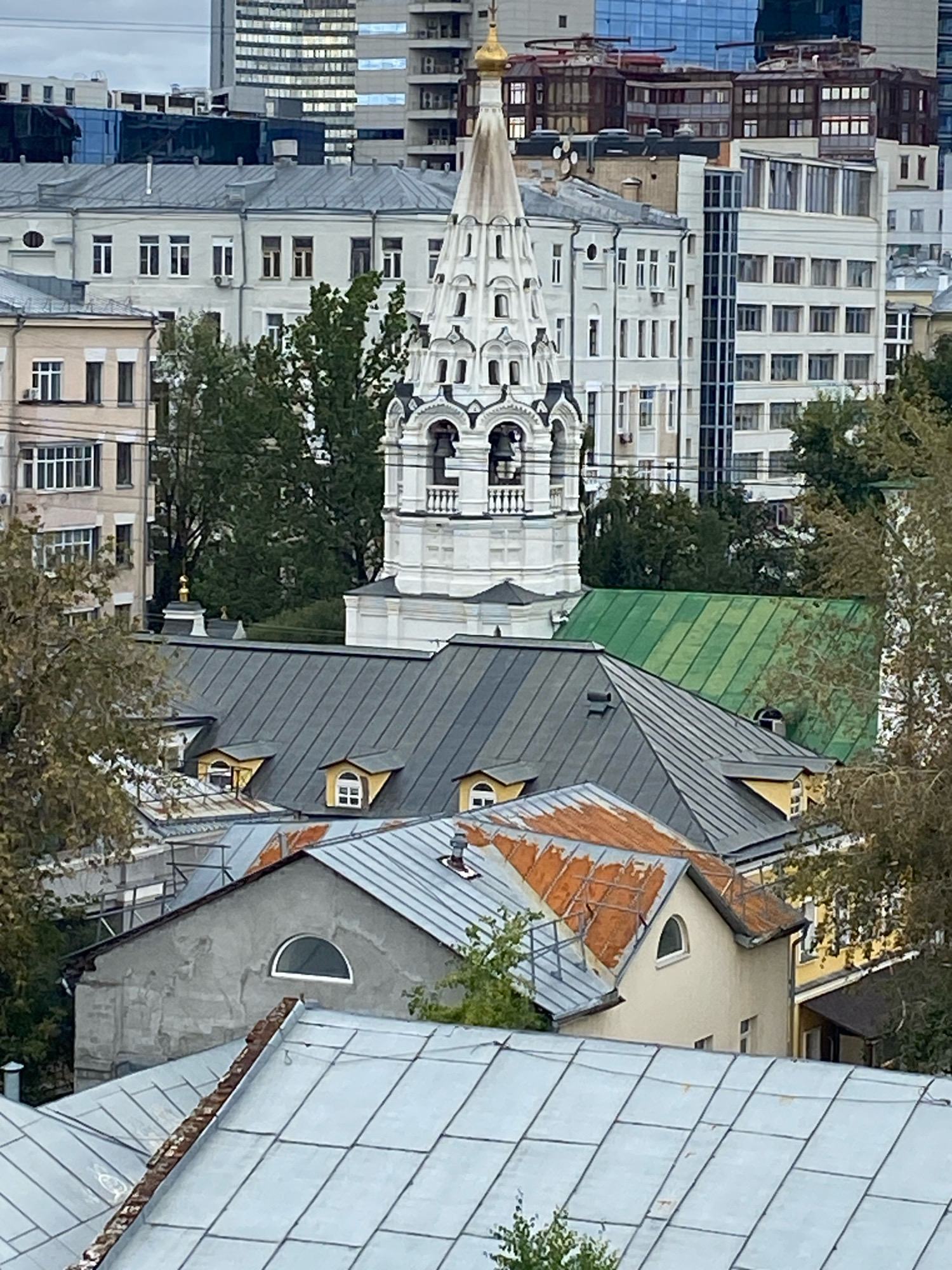The image displays an urban landscape in Moscow, Russia, characterized by a dense collection of buildings. In the foreground, several rooftops are visible, constructed from various materials including metal sheeting and weathered, possibly rusted, metal. One building features a grey, stucco exterior with a distinctive arched window.
Dominating the mid-ground is a white, ornate church tower, likely an Orthodox Christian church, with a golden dome. This structure is surrounded by other buildings of varying architectural styles and heights. To the right of the church tower, a brightly colored green roof is visible, contrasting with the muted tones of other structures.
In the background, modern high-rise buildings with numerous windows punctuate the skyline, indicative of a bustling city environment. Trees, some with autumnal foliage, are interspersed among the buildings, suggesting the presence of green spaces within the urban setting. The sky is overcast, contributing to the subdued lighting of the scene. No people are visible in the image.
