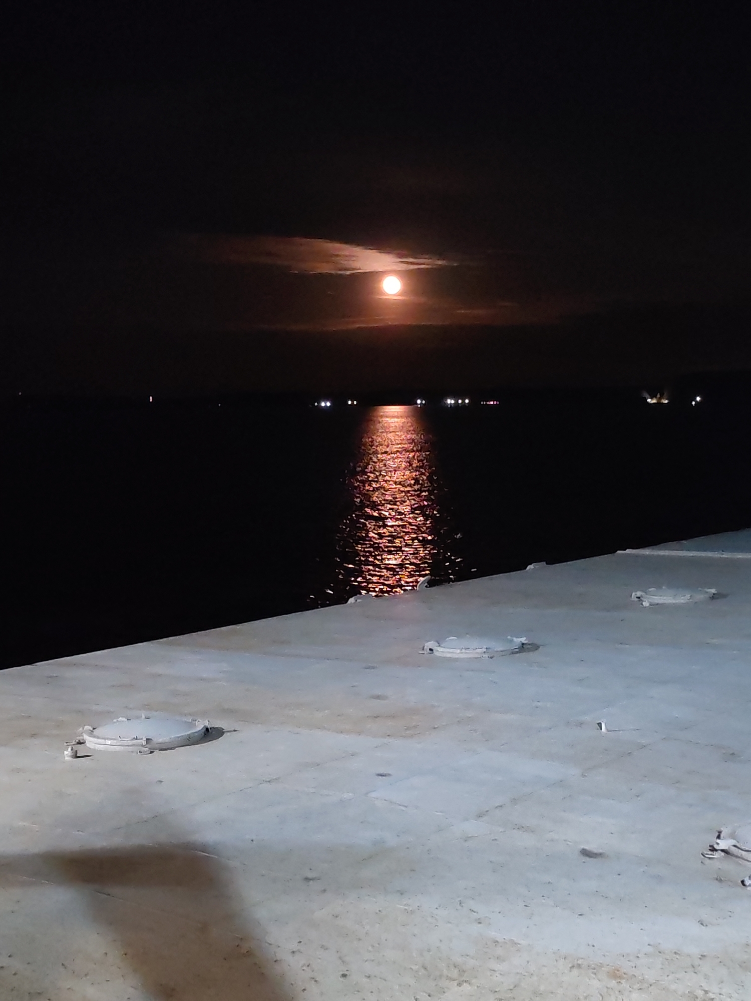 A nighttime view from a concrete platform looking out over dark water. In the sky, a full moon shines through a layer of clouds, casting a bright reflection on the water's surface. Distant lights twinkle on the horizon. The concrete platform in the foreground has several circular hatches embedded in its surface. The location is Unknown, Unknown.