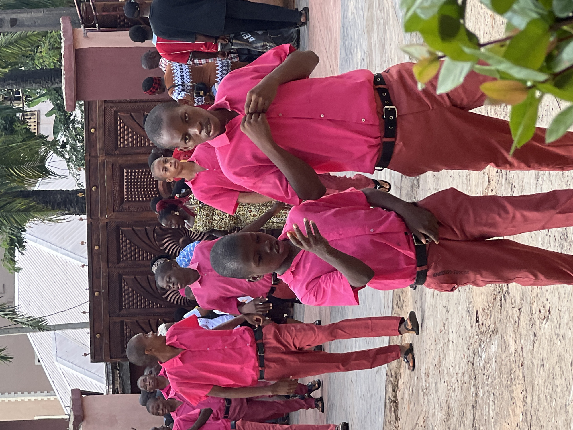 The image captures a scene in Nnewi, Nigeria, featuring a group of young males dressed in matching bright pink shirts and reddish-brown trousers. The individuals are walking outdoors on a paved surface, possibly a school compound or a public event area, given the presence of an ornate wooden gate and a building with a white corrugated roof in the background.
In the foreground, two boys are prominently featured. The boy on the right, facing forward, is buttoning his pink shirt. He wears a black belt with a gold buckle. To his left, another boy, also in pink and reddish-brown attire, is looking to his side and appears to be gesturing with his right hand. Other individuals in similar pink and reddish-brown outfits are visible in the midground and background, suggesting they are part of a unified group or attending the same event.
The environment appears to be warm and possibly sunny, with palm trees visible. The overall impression is one of an organized gathering, possibly a school event or a ceremonial procession. The wooden gate in the background suggests a traditional or culturally significant setting.