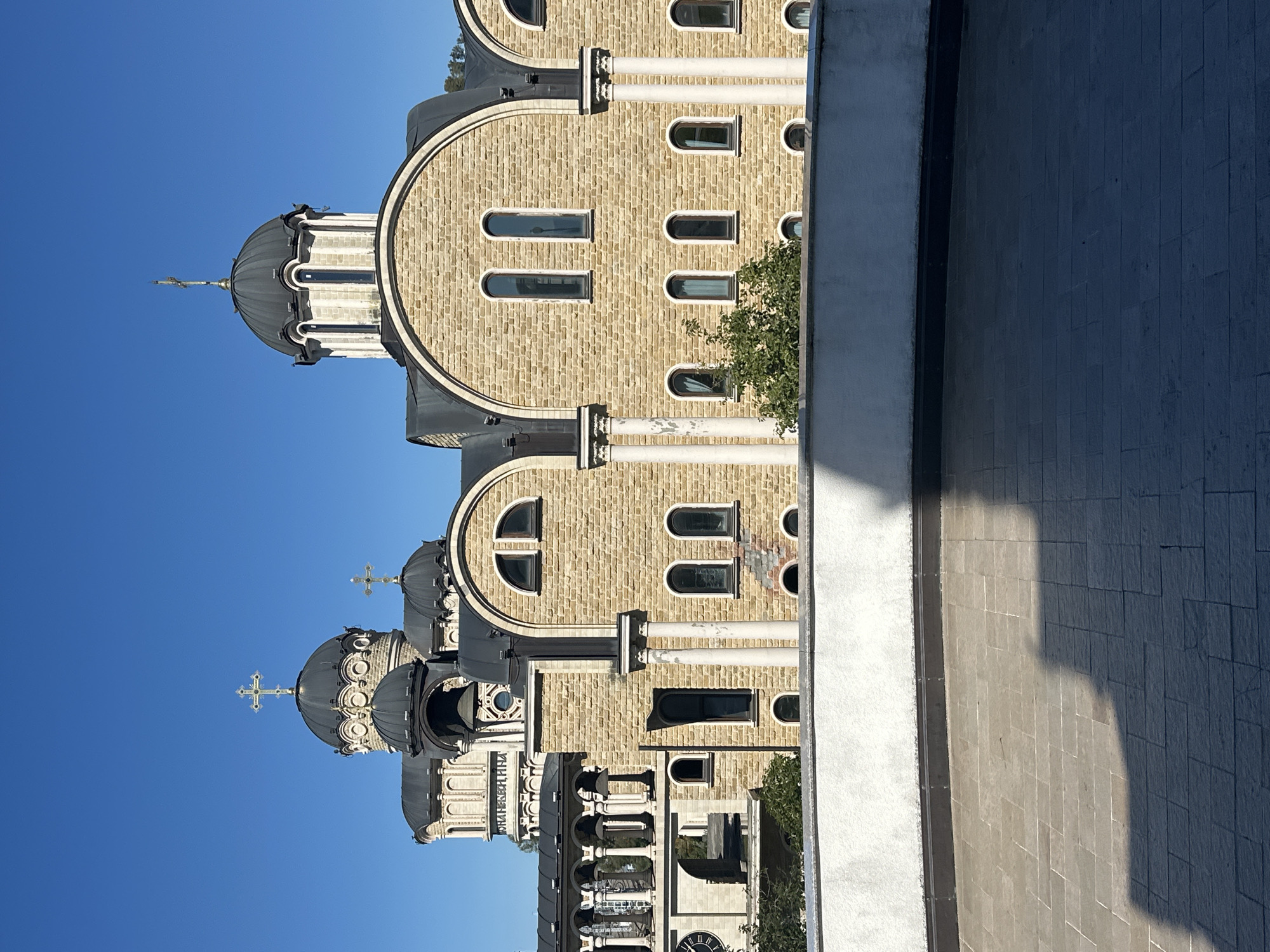 The image shows the administrative building of the Sirius Educational Center, located in the city of Sochi, Russia. The building features a light-colored stone facade with several domes, characteristic of orthodox architecture. The domes are topped with crosses, and arched windows are visible on the facade. In the foreground, there is a paved area with a white wall in front of it, casting a shadow. The sky is clear and blue.