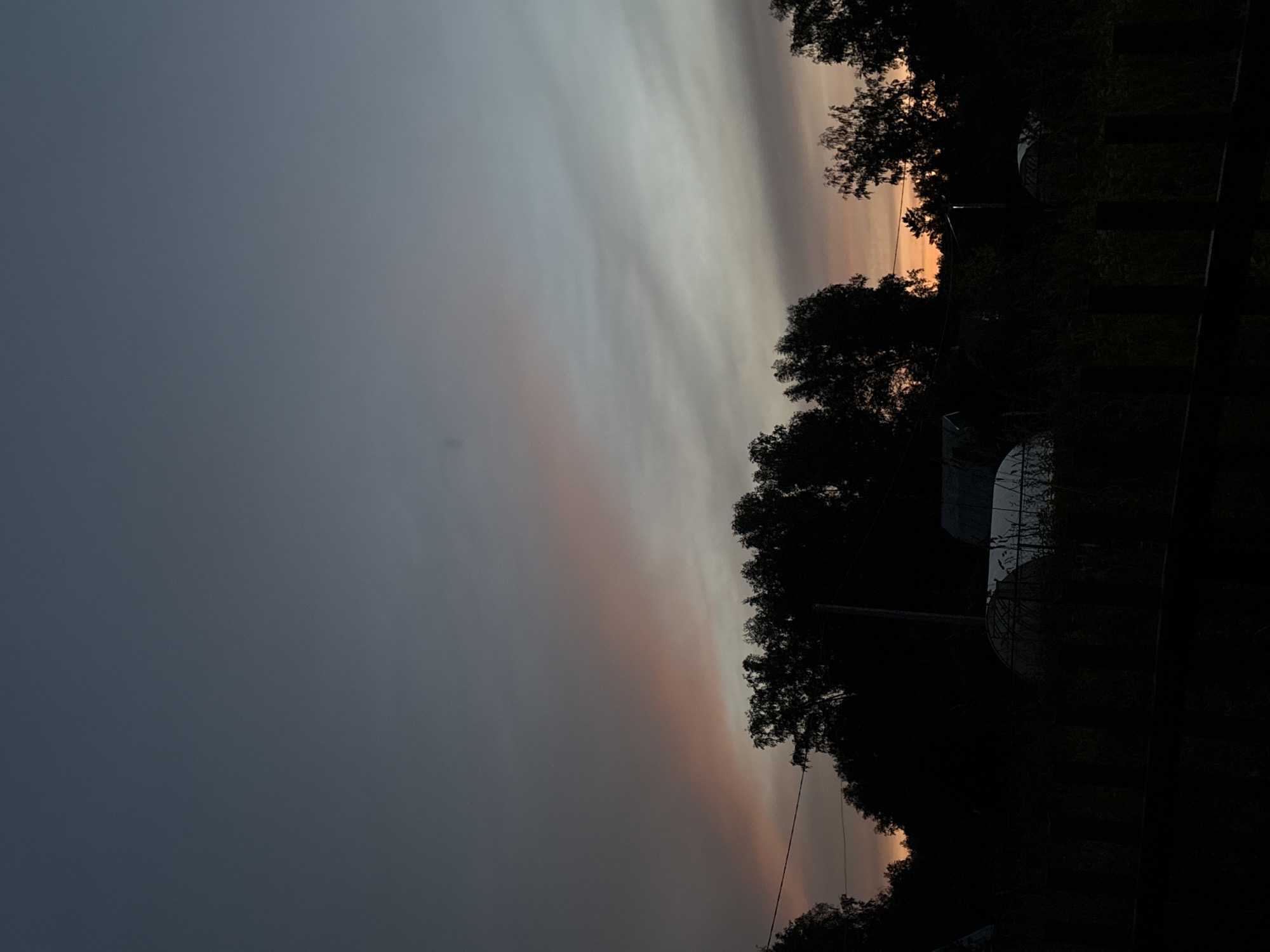 The image captures a dusky sky with hues of gray, white, and a faint blush of orange from a setting sun. Silhouetted trees form a dark, dense line along the right side of the frame, partially obscuring some dome-shaped structures and a fence. The sky is the dominant subject, with wispy clouds stretching horizontally. The overall mood is serene and contemplative, suggesting an evening in a rural or semi-rural setting. The low light conditions create a sense of peaceful transition from day to night. There is no visible text in the image. The location is identified as Dubrovino, Russia.