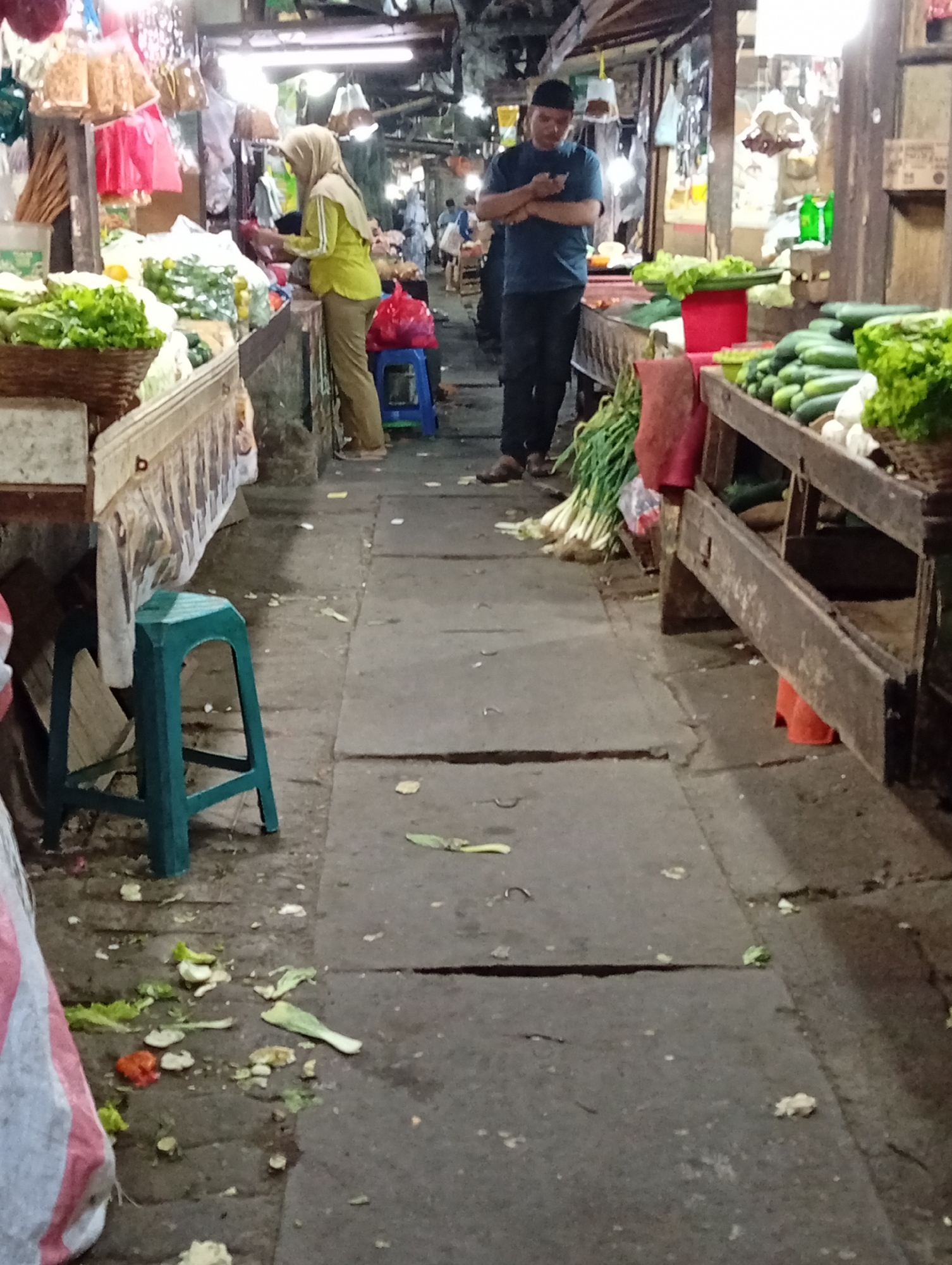 In Larangan, Indonesia, a bustling marketplace is captured in this image. The scene unfolds within an indoor market, characterized by rows of vendors selling produce. In the foreground, a green plastic stool stands on a textured concrete floor littered with vegetable scraps and debris. To the left, a stall displays an assortment of vegetables in woven baskets, including lettuce and other leafy greens. Further into the market, a person wearing a hijab and a yellow shirt and brown pants is seen attending to a display of produce. Opposite them, a man in a blue t-shirt and dark pants stands with his hands crossed, looking at his phone. The market stalls are constructed from wood, with tables laden with various vegetables such as cucumbers and scallions. Bags of produce and red plastic bags are visible. The lighting suggests it is either early morning or evening, with artificial lights illuminating the market.