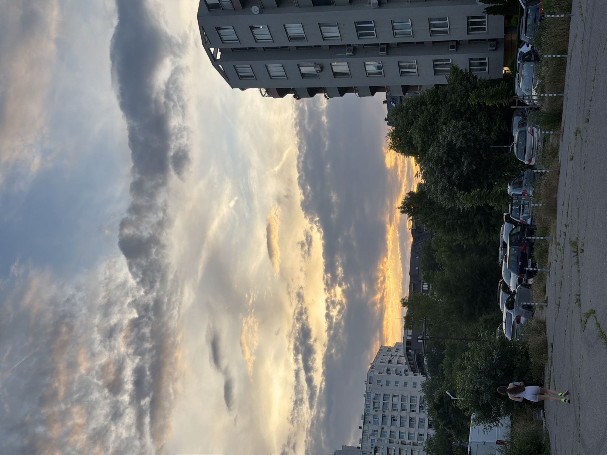 The image depicts a street-level view of an urban setting in Sofia, Bulgaria, at sunset. The sky is filled with dramatic clouds, illuminated by the setting sun, creating a mix of orange, yellow, and grey hues. In the foreground, a woman with long brown hair is seen from behind, wearing a light grey athletic outfit and bright yellow socks, jogging away from the camera. She is holding a light blue object, possibly a phone or a water bottle. She is running on a cracked asphalt surface, which appears to be a parking lot or an abandoned road.
To the left of the frame, a large, multi-story apartment building is visible, with numerous windows and balconies. Further back, another residential building, also appearing to be an apartment block, is partially obscured by trees. In the middle ground, a dense line of green trees separates the foreground from the apartment buildings. Beyond the trees and parking lot, a row of parked cars is visible, including a silver sedan and a dark hatchback.
To the right of the image, a tall, modern apartment building with a distinctive curved facade and numerous windows stands prominently. Several air conditioning units are mounted on its exterior. The overall impression is of a residential area in Sofia during the evening, with the sky being the most striking element.