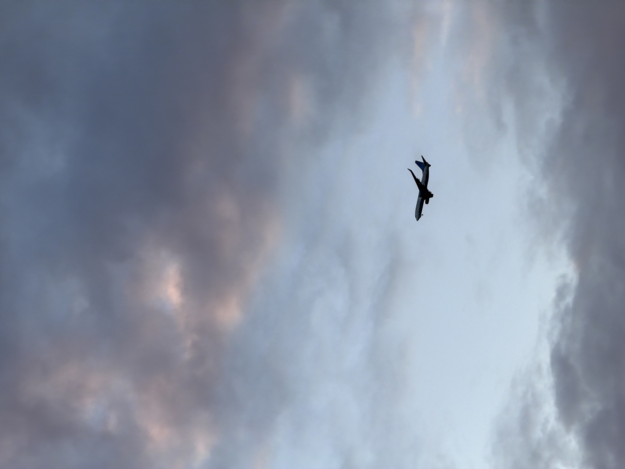 The image captures a commercial airplane in sharp silhouette against a cloudy sky. The aircraft is positioned in the upper right portion of the frame, depicted from below, with its wings and fuselage angled, suggesting it is banking or turning. Its dark form stands out against the brighter parts of the sky.
The sky is largely filled with clouds, presenting a dynamic range of tones. Dense, dark grey clouds occupy the left and lower right sections, transitioning to lighter blue-grey and almost white areas towards the center and upper right. Subtle hints of pinkish-orange light are visible within some of the darker cloud formations, suggesting either the diffuse light of a heavily overcast day or the early stages of a sunset or sunrise reflecting on the clouds.
No people or discernible text are visible in the media. The scene depicts an aerial view, emphasizing the vastness of the sky and the solitary passage of the aircraft. Given the context, this aviation scene unfolds over Perm, Russia, likely during an overcast day or at dusk/dawn.