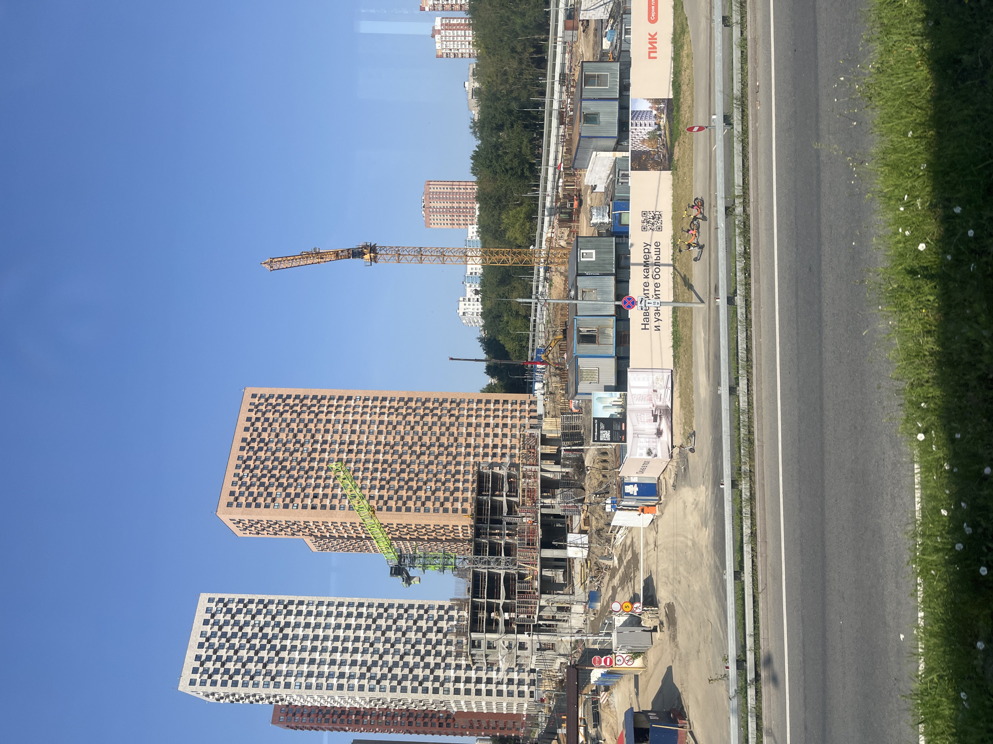 The image depicts a construction site in Moscow, Russia, with several high-rise buildings under construction. In the foreground, a highway with a metal guardrail separates the viewer from the construction area. The construction site features a large yellow crane, a green construction crane, and various modular buildings. In the background, a line of trees separates the construction site from more residential buildings, some of which are also under construction or recently completed. The sky is clear and blue. There are no people clearly visible in the scene, though there are two electric scooters parked near a fence with advertising banners. The overall impression is one of ongoing urban development.