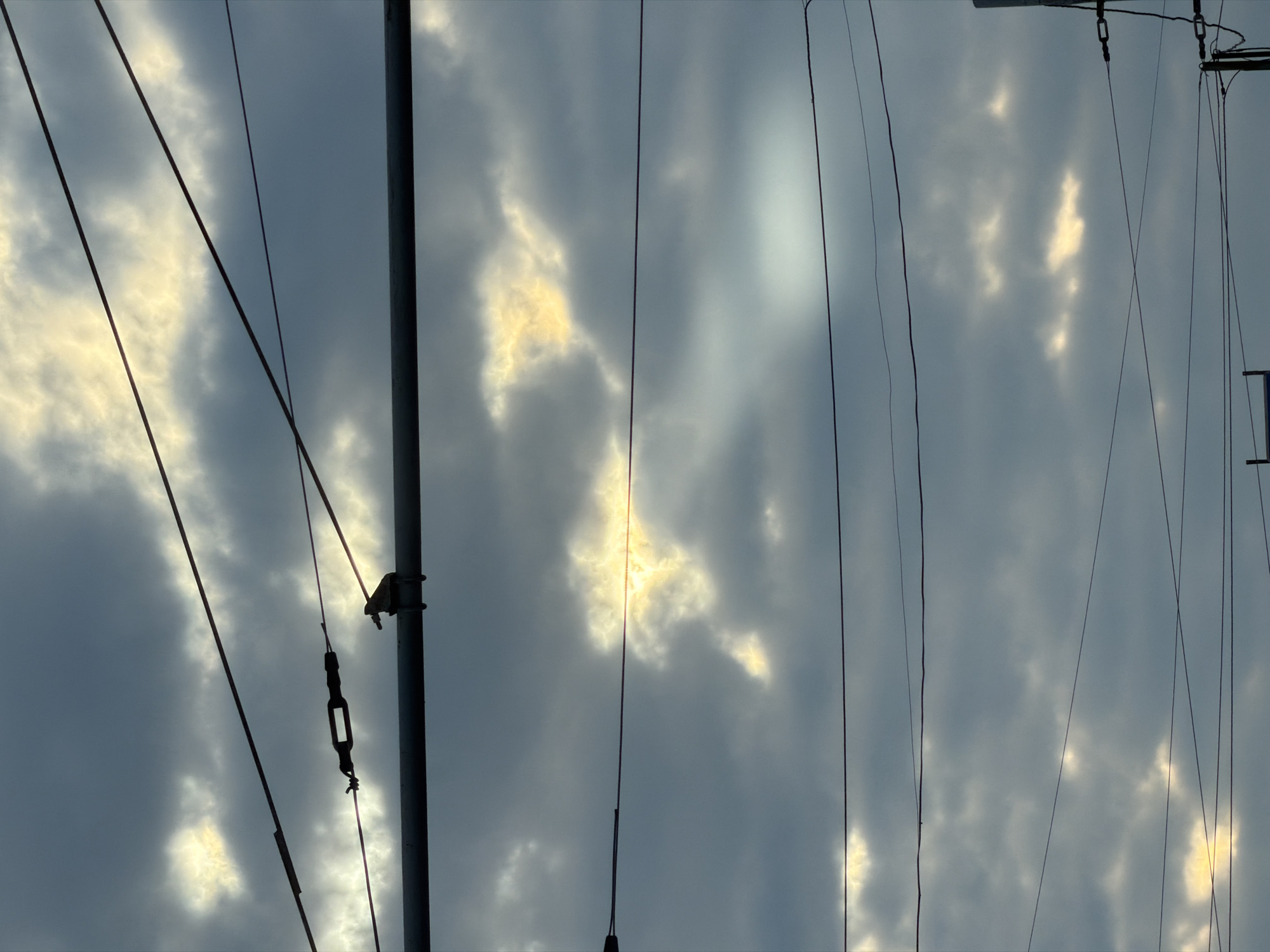 The image shows a view looking up at a cloudy sky. The sky is gray with breaks in the clouds where sunlight is visible. In the foreground, there are vertical and angled wires and a pole, possibly electrical or communication lines. The overall impression is one of overcast weather with the built environment intruding into the sky. There are no people visible. There is no information to confirm the location of the photo, but it has been requested that it be in Izhevsk, Russia.