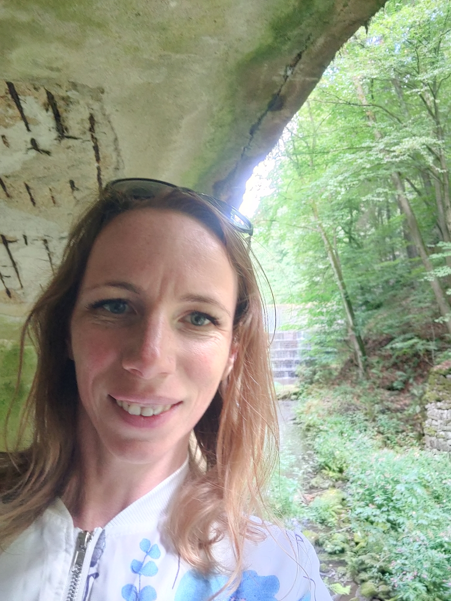 In a selfie taken at the Polenztal valley in Lohmen, Germany, a fair-skinned woman with shoulder-length light brown hair and blue eyes smiles at the camera. She wears sunglasses perched on her head and a white zippered jacket adorned with a blue floral pattern. The woman is positioned in the foreground, partially obscured by an overhead rock overhang with visible rebar, suggesting an old structure or cave. Behind her, a lush, green forest fills the background. A stone staircase descends into the valley to her right, with a stream and dense vegetation visible at the bottom. The lighting suggests it is daytime, with dappled sunlight filtering through the trees.