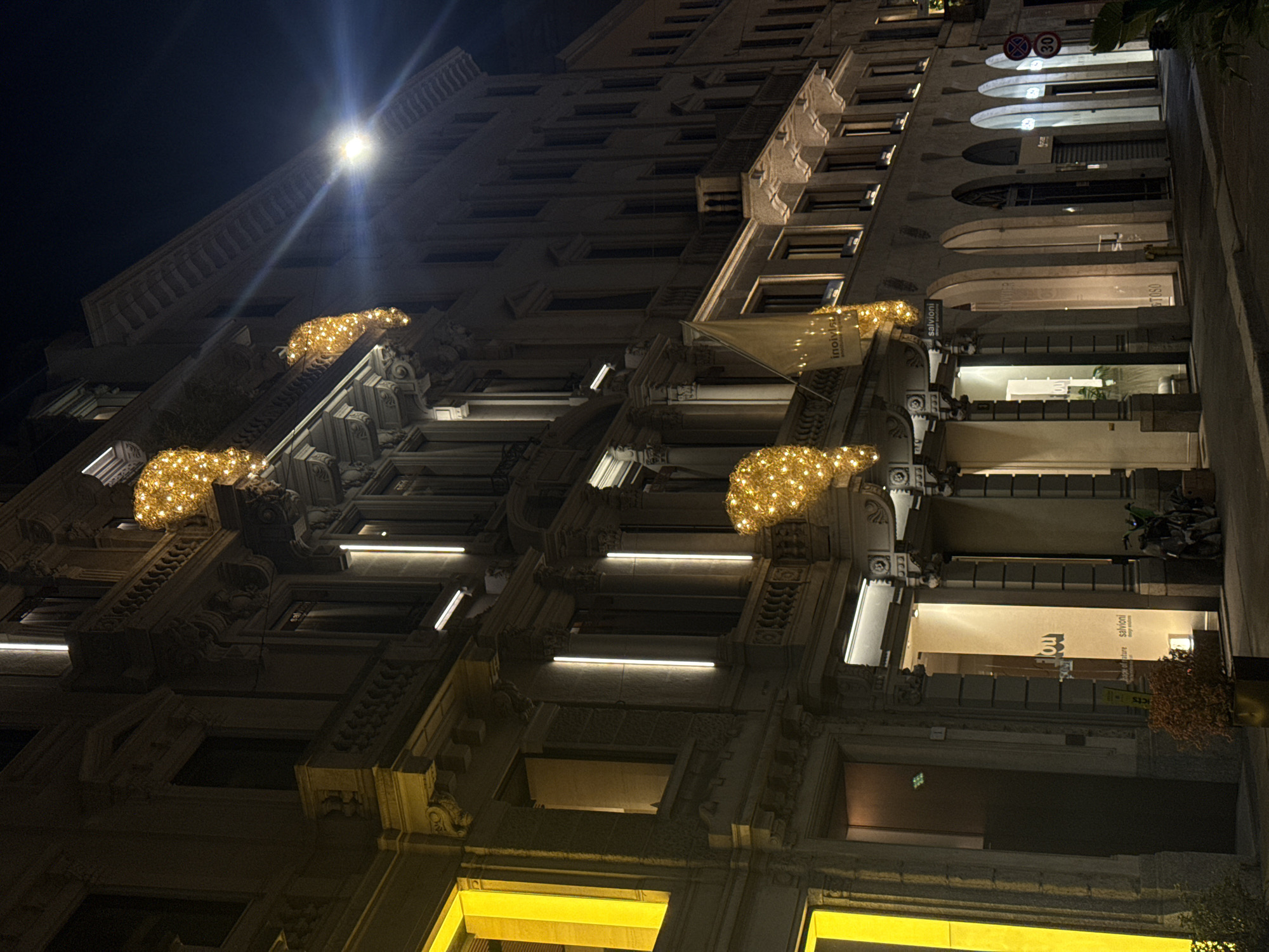 The image captures a tall, ornate building at night in Milan, Italy, viewed from a low angle, looking upwards. The multi-story structure is illuminated by various artificial light sources, creating a festive and elegant urban scene.
The building's facade is constructed from what appears to be light-colored stone or concrete, featuring classical architectural elements such as decorative corbels, balustrades, and arched window alcoves. Horizontal linear lights, emitting a bright white glow, illuminate the undersides of balconies and window frames, highlighting the building's intricate details.
Prominently displayed on the facade are several large, golden light installations shaped like crouching animals, possibly bears or stylized lions, adding a whimsical and festive touch. A bright, starburst-like street light or spotlight is visible high up and to the left, casting a strong glare.
Inside one of the larger ground-level shop windows, warm interior lighting reveals several signs. Visible text includes "involu" (partially inverted on a banner and inside the shop) and "salvioni" (also on a banner and inside a shop window, next to smaller text that appears to say "box of store"). In an upper arched alcove, what looks like several parked scooters or motorbikes can be discerned. High on the building, a red circular road sign with "30" inside and another red circular sign with a diagonal line (possibly indicating 'no entry' or 'no stopping') are visible. Green foliage from nearby trees is also visible in the upper right and lower right corners of the frame.
The overall impression is one of a sophisticated city street decorated for the evening or a holiday season, with no people visible, lending a calm ambiance.