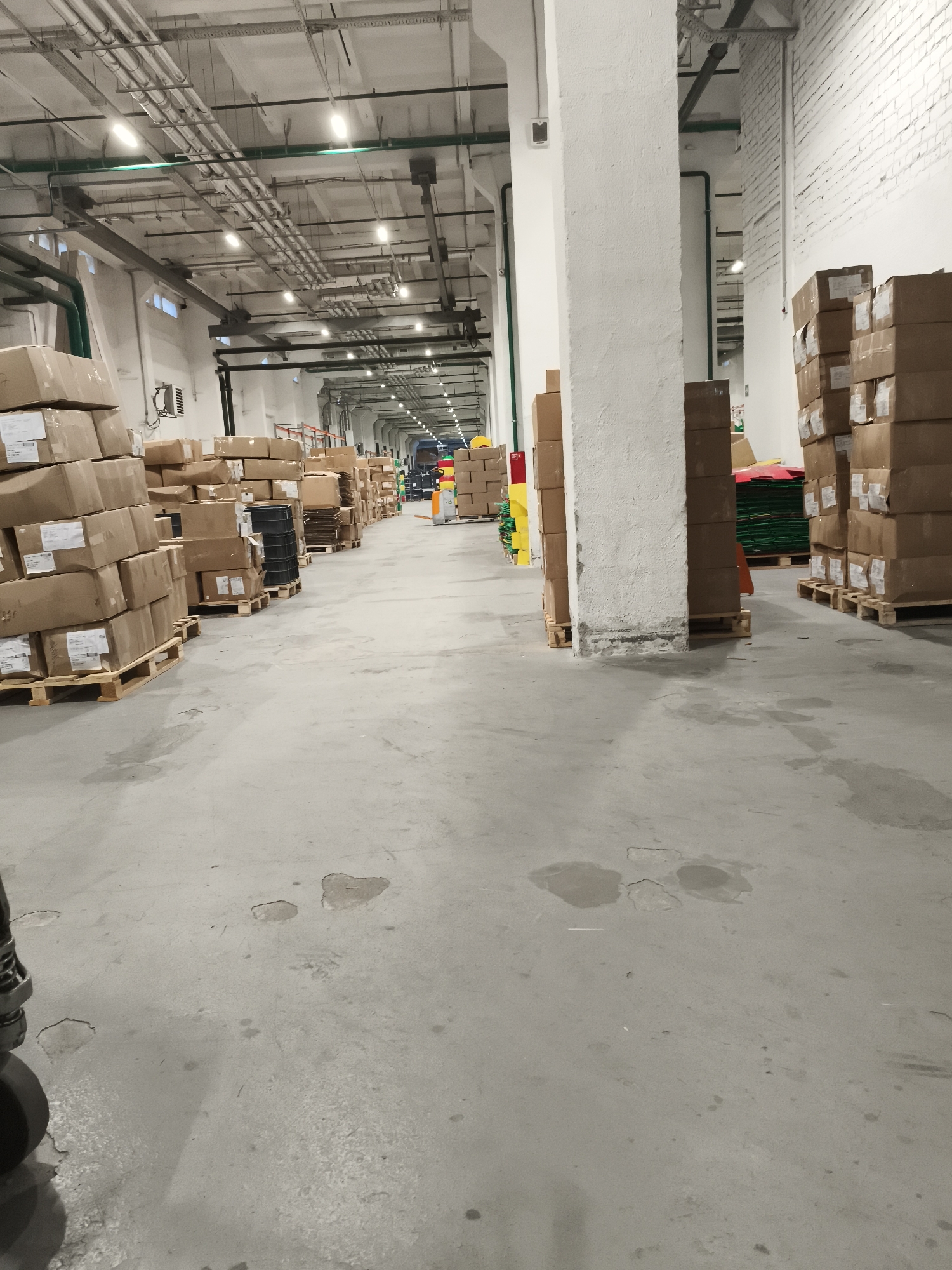 The image depicts the interior of a large warehouse, likely a distribution center. The scene is characterized by rows of stacked cardboard boxes on wooden pallets, occupying a significant portion of the visible floor space. These boxes vary in size and are arranged in aisles, suggesting an organized storage system. The warehouse features a high ceiling with exposed industrial lighting and a network of pipes and conduits running across it. Structural pillars are visible throughout the space, supporting the roof and dividing the areas. The floor is concrete and appears somewhat worn, with some stains and minor damage in places. In the background, additional stacks of boxes and some brightly colored items, possibly protective equipment or inventory, can be observed, extending deeper into the facility. The overall impression is one of an active logistics or storage environment. Given the absence of any specific identifying markers within the image, it is not possible to determine the exact location in the city of Barysaw, Belarus.