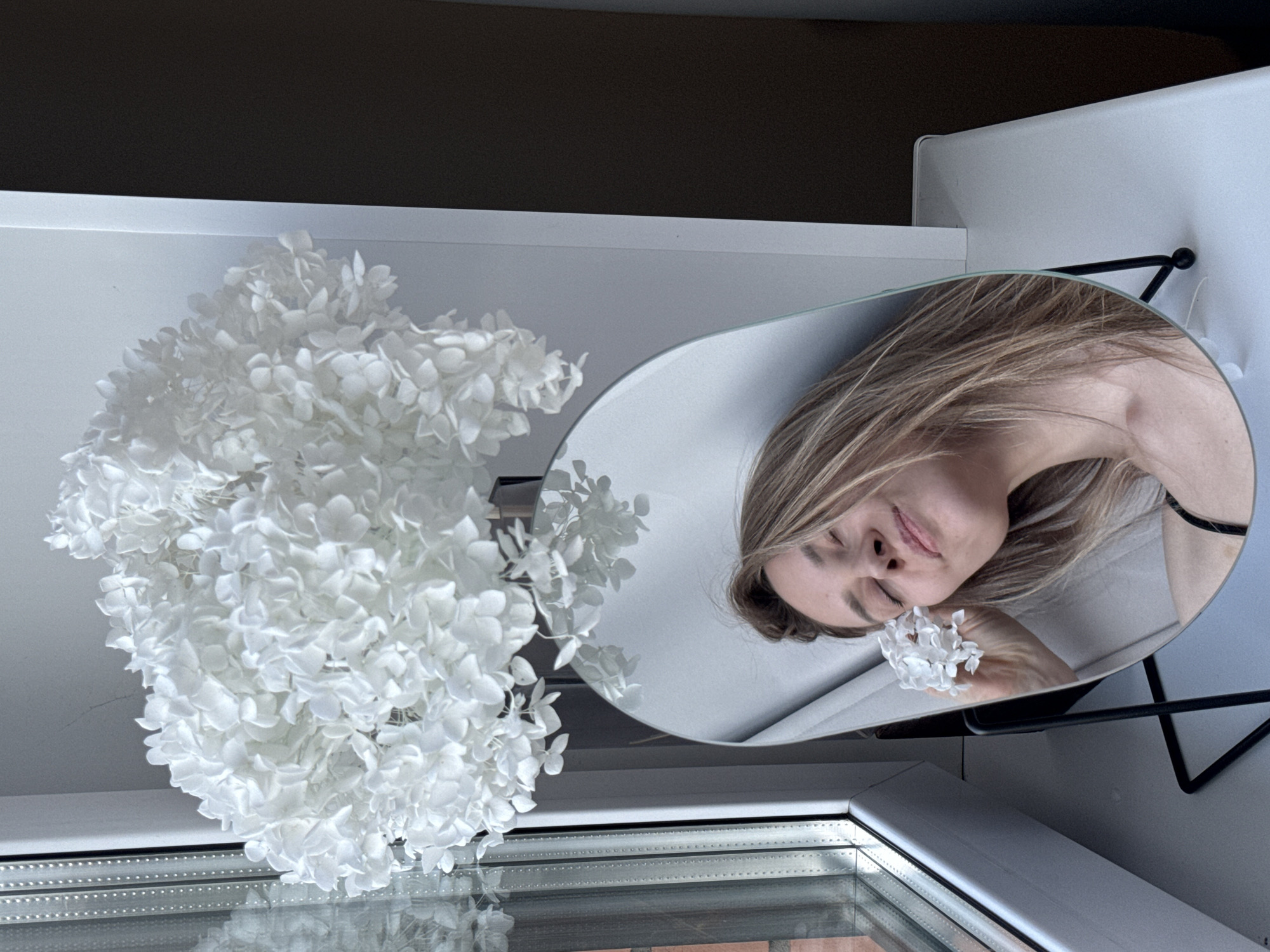 A woman with light brown hair is reflected in a circular mirror next to a large bouquet of white flowers. She is holding a small cluster of white flowers near her face. The scene appears to be indoors, potentially near a window, with a glimpse of a window frame and possibly a brick exterior visible. No specific details indicate this is Chaikovsky, Russia.