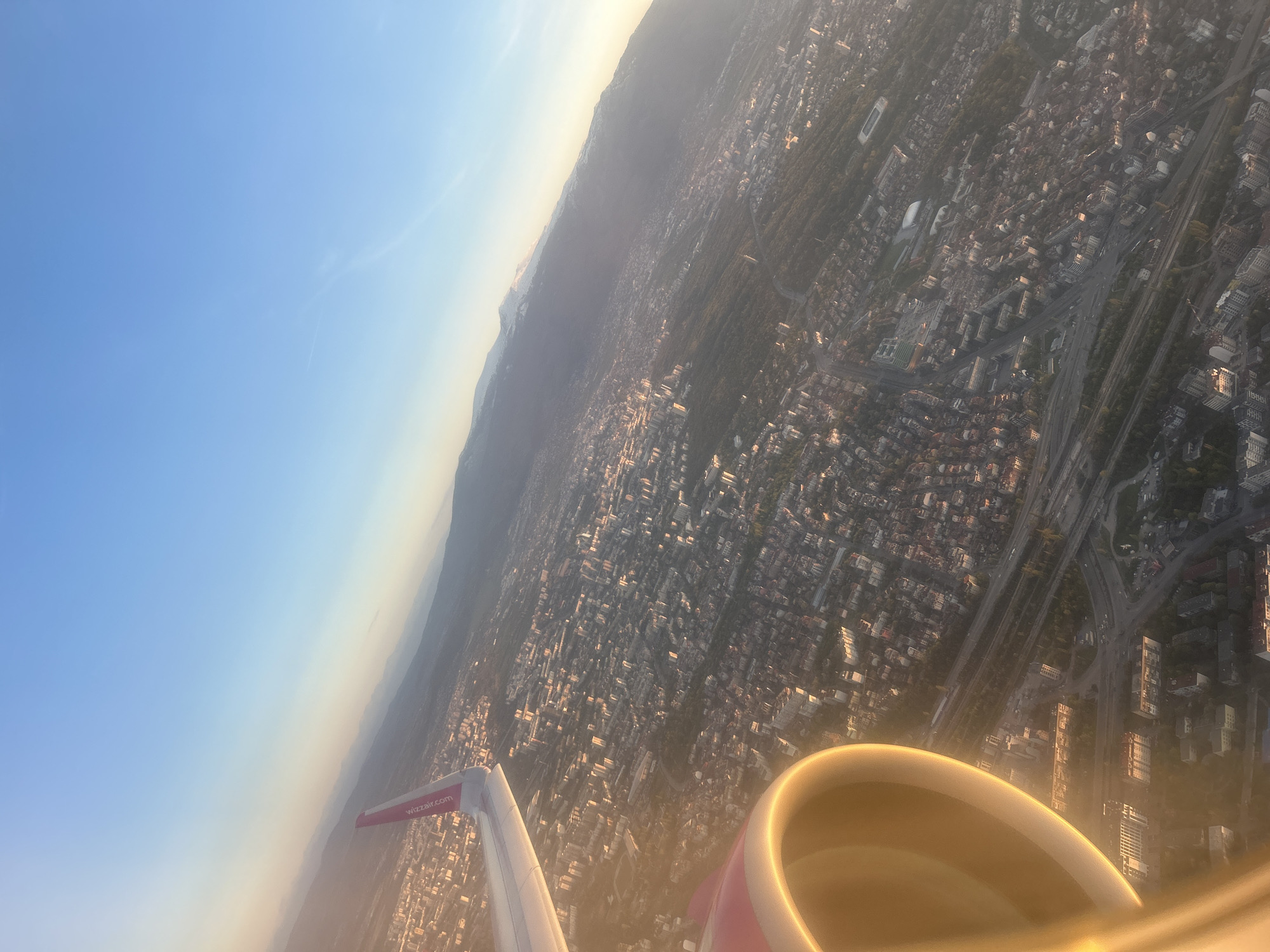 The image, taken from an airplane, shows the city of Sofia, Bulgaria. The plane's wing and engine are visible in the foreground, indicating the viewpoint is from inside the aircraft. Below, a dense urban landscape is visible with buildings, roads, and greenery. In the background, a mountain range is visible. The lighting suggests either sunrise or sunset, casting a warm glow over the city.