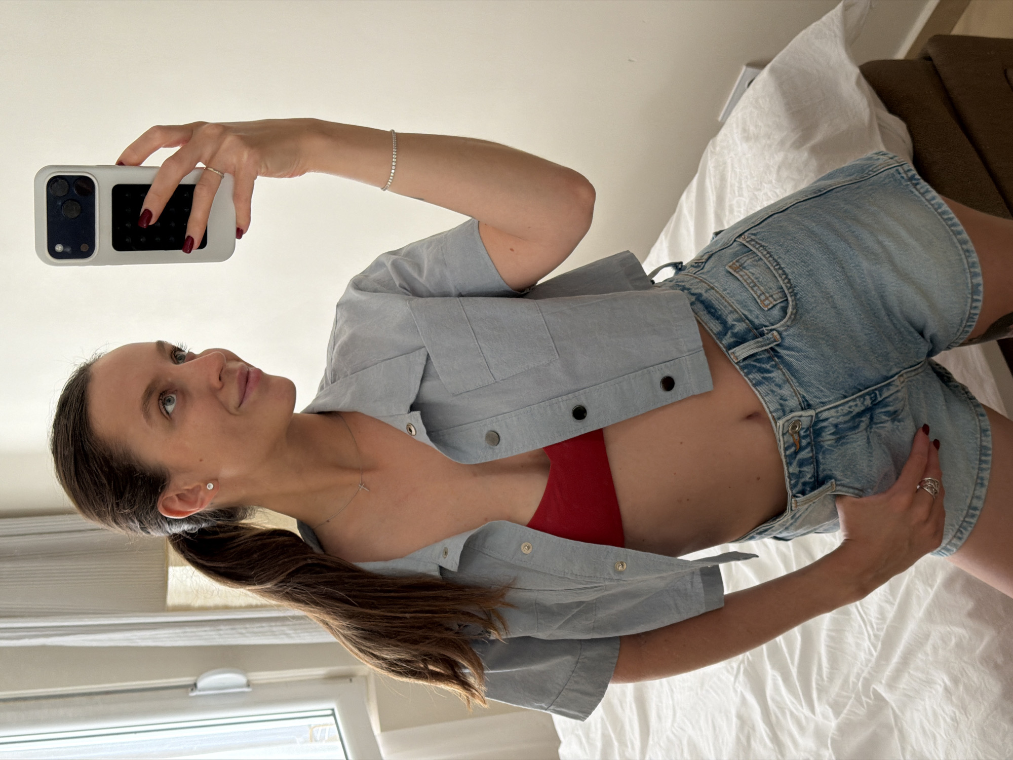 A woman takes a selfie with her phone on a bed in a room with a window. She wears denim shorts, a light blue button-down shirt, and a red top underneath. The phone has a white case with black camera lenses and buttons. She wears rings on both hands and a bracelet on her right wrist, and her nails are painted dark red. Her brown hair is pulled back in a ponytail. A window and white curtain are visible in the background. The location is not apparent from the image, and there is no visible evidence to suggest that the scene is in Budva, Montenegro.