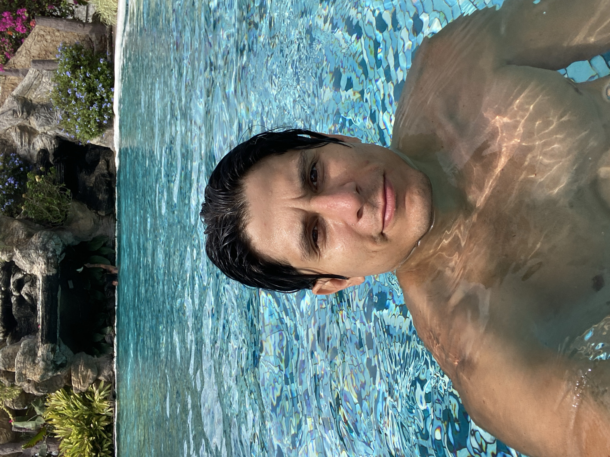 A man is partially submerged in a swimming pool, taking what appears to be a selfie, looking directly at the camera with a neutral, relaxed expression. His upper torso and head are visible above the waterline. The clear blue water reflects bright sunlight, creating shimmering patterns on his skin and the mosaic tiles of the pool floor. His hair is dark and appears wet.
The swimming pool has a naturalistic edge on the left, featuring stacked rock formations and lush green vegetation. Various flowering plants are visible, including vibrant purple bougainvillea and smaller blue flowers, suggesting a tropical or resort-like environment. The background indicates a sunny daytime setting, with the bright light reflecting off the water. The overall scene conveys a sense of leisure and relaxation. There is no visible text in the image. Given the context, this tranquil setting is likely in Vallecito, Venezuela. The image is oriented sideways, with the man's head positioned towards the top of the frame.