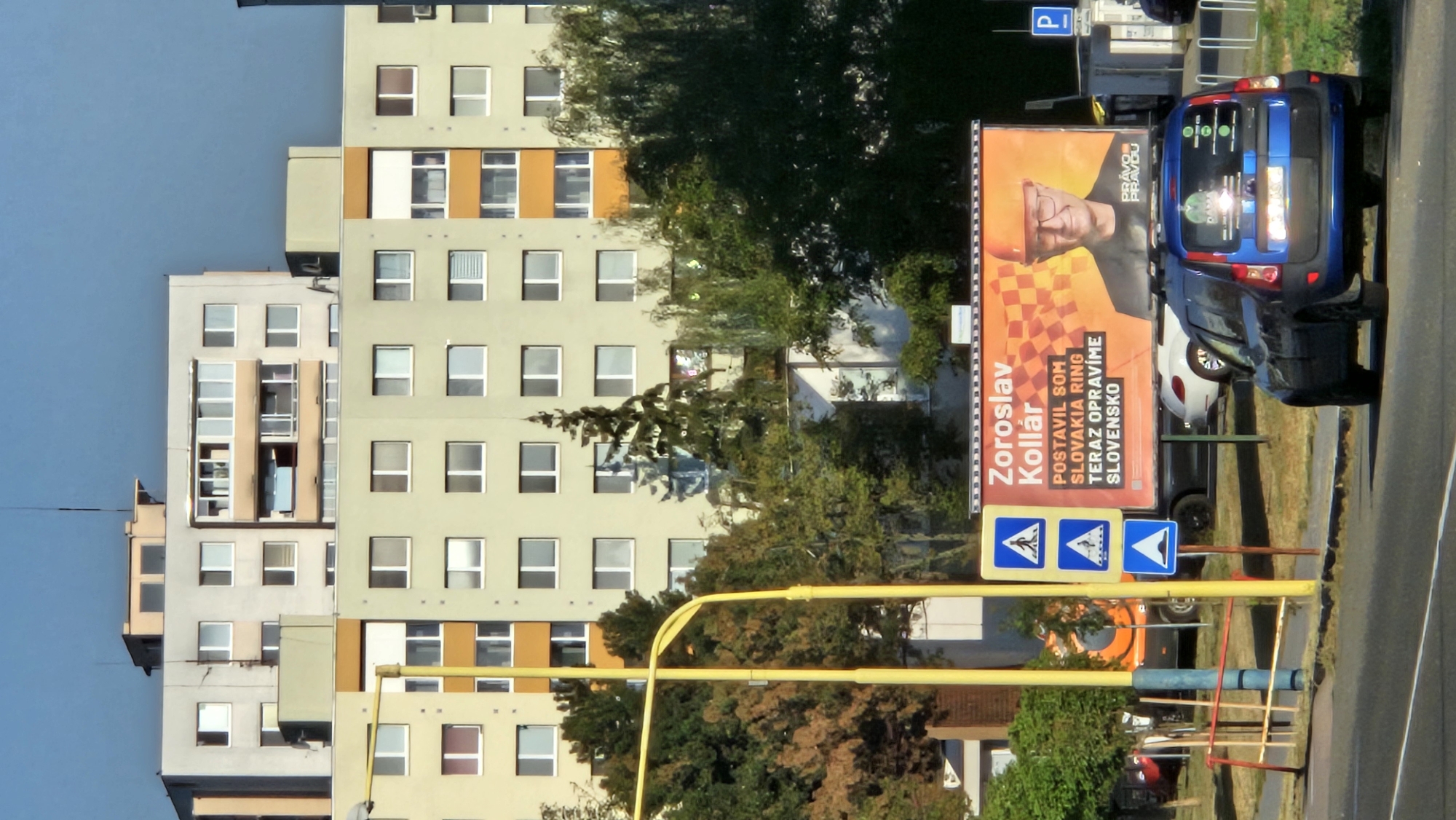 The image captures a street scene in Prešov, Slovakia, dominated by a large, modern apartment building in the background and a billboard in the mid-ground. The apartment building is light-colored with numerous windows and several balconies, some of which have visible occupants or items. In front of the building, a lush green tree stands tall, partially obscuring the view of the structure.
The billboard features a portrait of a man wearing an orange hard hat and glasses. Text in Slovak is visible, including "Zoroslav Kollár" and "POSTAVIL SOM SLOVAKIA RING TERAZ OPRAVÍME SLOVENSKO," suggesting a political or public service message. To the left of the billboard are three blue pedestrian crossing signs stacked vertically.
In the foreground, a dark blue car is parked on the right side of the road, facing away from the viewer. The road itself is paved and marked with white lines. A yellow pole, part of a street light or traffic signal, is prominently positioned to the left of the billboard, along with a smaller blue pole. Some fencing and other vehicles are also visible in the background, contributing to the urban environment. The sky is a clear, bright blue.
