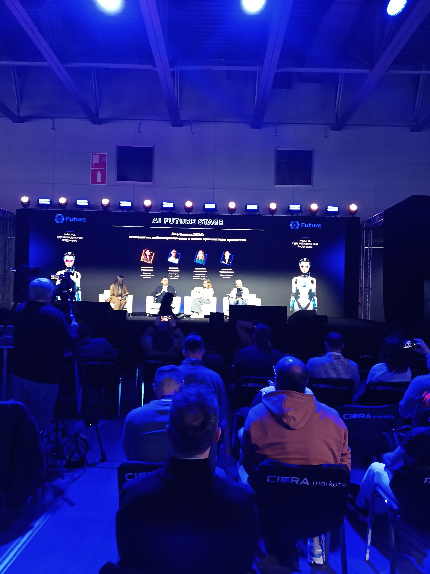 Here is a description of the image:
The image shows a conference or presentation on AI (Artificial Intelligence) called "AI FUTURE STAGE", which is part of the "Future" event. The setting appears to be a large hall, with rows of chairs filled with an audience. Many chair backs have the label “CIERA markets.” Onstage, there is a large screen displaying the event logo and details about the speakers and the topic being discussed: "Al в банках 2026: Новая архитектура процессов" (AI in banks 2026: New process architecture). Four speakers are seated on white chairs in front of the screen, along with two human-like robots standing on either side of the stage. A videographer is positioned on the left side of the frame, filming the event. The lighting is predominantly blue. Based on the Cyrillic text, the location is most likely in a Russian-speaking region, possibly Khimki, Russia.