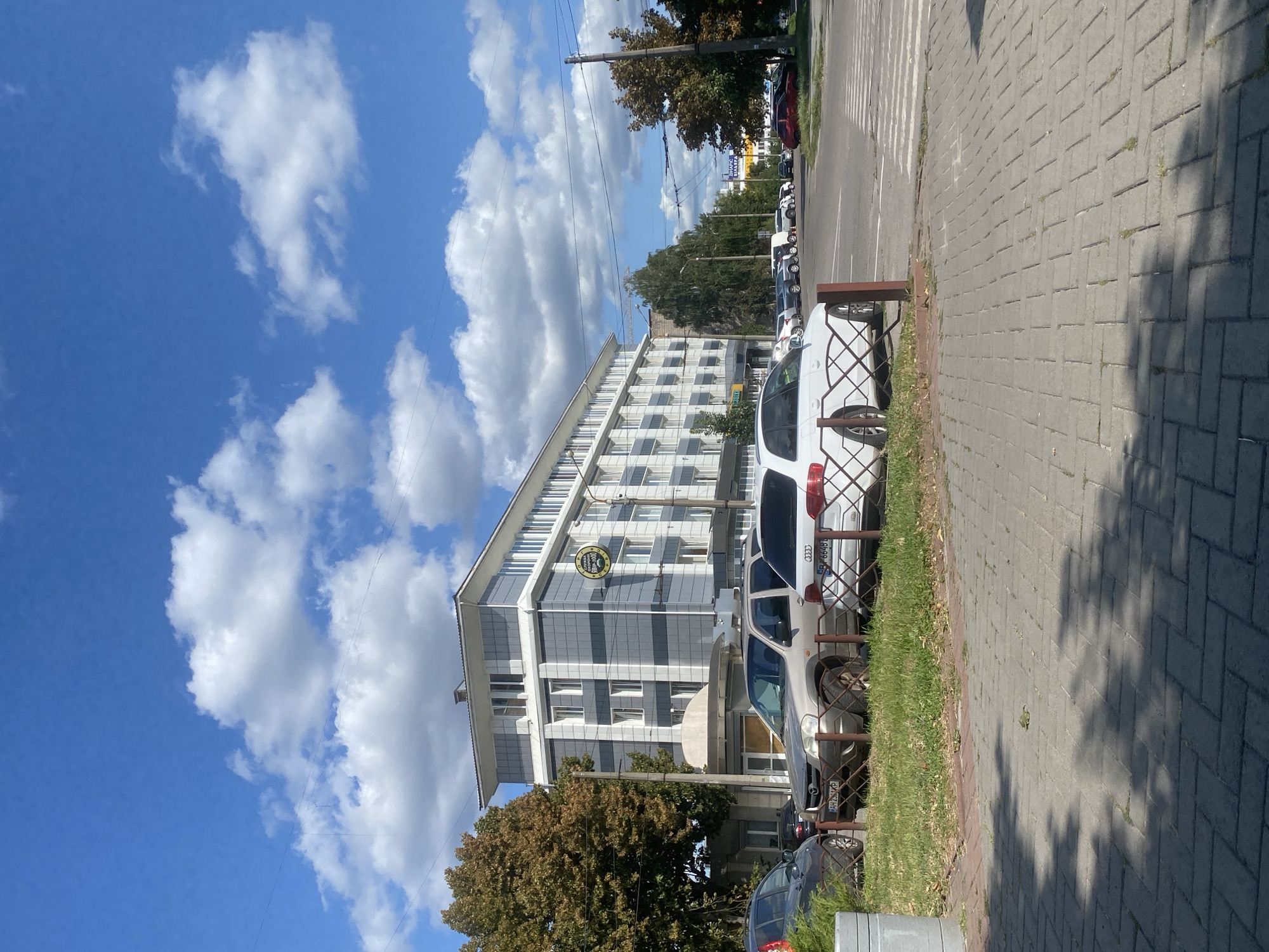 The image depicts the exterior of a modern, multi-story building with a grey and white facade, located in the city of Poltava, Ukraine. The building appears to be a hotel or office complex, as indicated by a circular sign with "КНИГА 3000" and a graphic of an open book, suggesting a hotel or book-related business. In front of the building, several cars are parked in a lot. These include a silver SUV, a white Audi station wagon, and a dark grey hatchback. The cars are parked on a grassy area bordered by a metal fence. The foreground shows a paved pedestrian walkway made of grey rectangular blocks, with a small green bush on the left. The sky is blue with scattered white cumulus clouds. Trees with autumnal foliage are visible on the left side of the building. The overall scene is a daytime view of an urban environment, likely a street in Poltava.