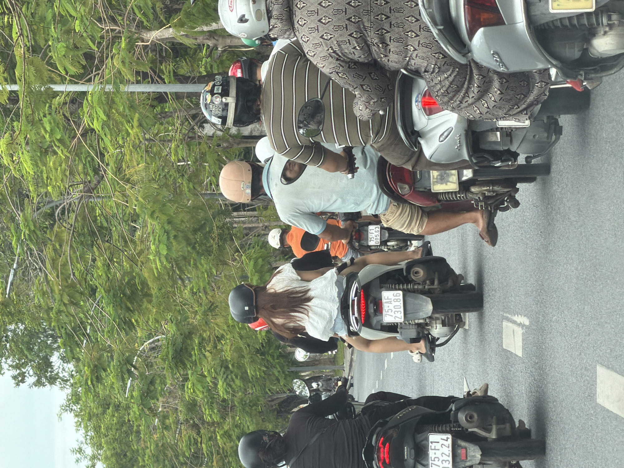 The image depicts a street scene in Phú Xuân Ward, Vietnam, with a line of motorcyclists traveling on a paved road bordered by lush green trees. The perspective is from behind the riders, focusing on their activity.
In the foreground, several individuals are seen riding scooters. The rider on the far left wears a black helmet and a black shirt. Next to them, another scooter rider is seen from the back, wearing a black helmet and a white top with denim shorts, their hair is long and brown. To their right, a rider wears a tan helmet and a light blue t-shirt with beige shorts. Further to the right, a person in a dark olive green and white striped shirt is on a scooter, wearing a black helmet adorned with stickers. The person to their right is wearing a light gray helmet and a patterned grey and black garment.
The scooters are various models and colors, including black, white, and silver. License plates are visible on some scooters, with the numbers "75-AF 230.86" clearly legible on one. The road surface shows white lane markings. The environment is characterized by dense green foliage lining the street, suggesting a well-treed urban area. The sky is overcast and light-colored. The overall impression is a snapshot of daily transportation in a Vietnamese city.