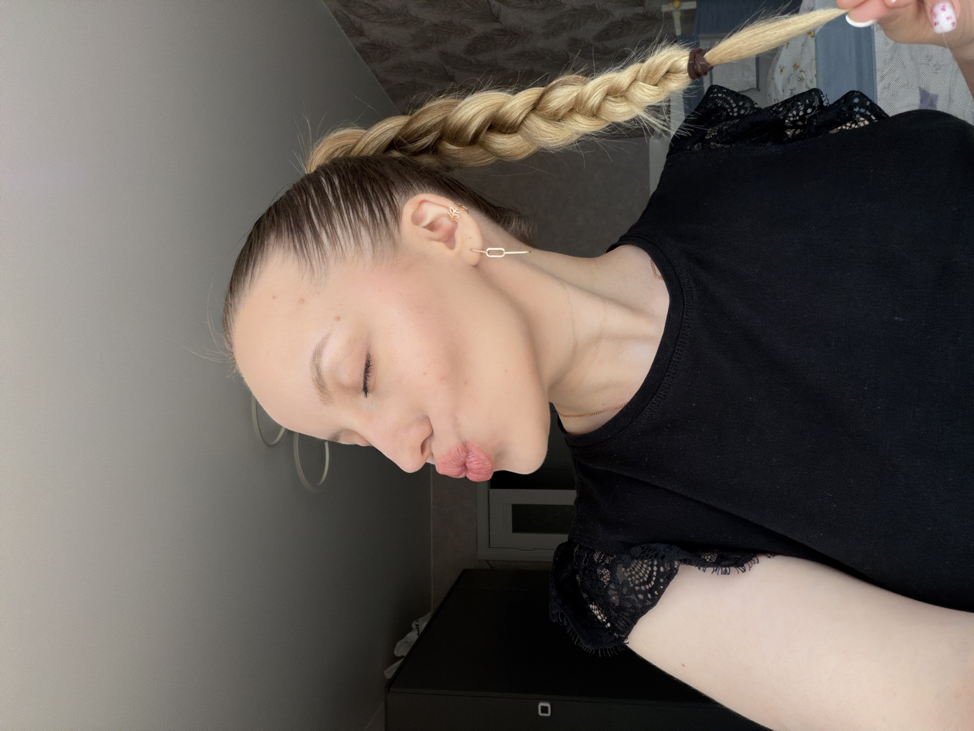 The scene depicts a solo portrait of a female subject, believed to be indoors in the city of Reshetnikovo, Russia. The subject, a fair-skinned individual, is shown in profile facing to the left, with her eyes closed and lips pursed in a kissing gesture. She has light blonde hair styled in a tight braid extending over her right shoulder, with darker roots visible. She wears a black short-sleeved top featuring lace detailing on the shoulders and sleeves. Accessories include a thin gold-colored chain necklace, a rectangular link earring in her right earlobe, and a gold-colored ear cuff spelling "R" on the helix of the same ear. Her right hand is visible in the lower right corner, gently holding the end of her braid, which is secured by a dark brown elastic band. The fingernails of this hand are painted a light color with small pink or red patterns. The background shows a dimly lit room with a dark wardrobe or cabinet on the left. The walls are covered with a light-colored, possibly textured wallpaper. A white door frame is partially visible in the center-right background. In the lower right, a portion of a bed with a blue and white patterned duvet or blanket is visible. The ceiling is grey with two circular light fixtures partially visible.