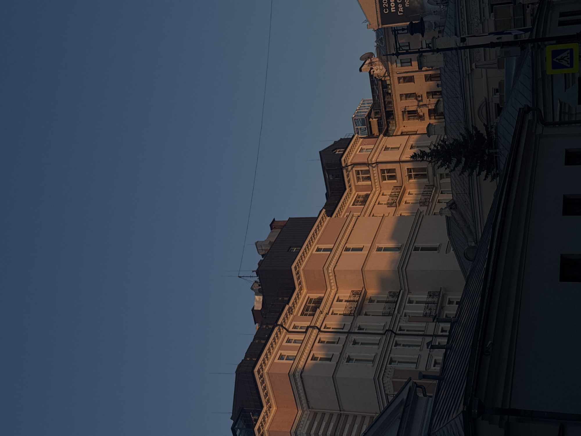 The image shows a street in Moscow, Russia, with several buildings visible. The buildings are multi-story structures with varying architectural styles and color schemes. The sky is a clear blue and some buildings are covered in golden light. A pedestrian crossing sign and utility lines are visible.