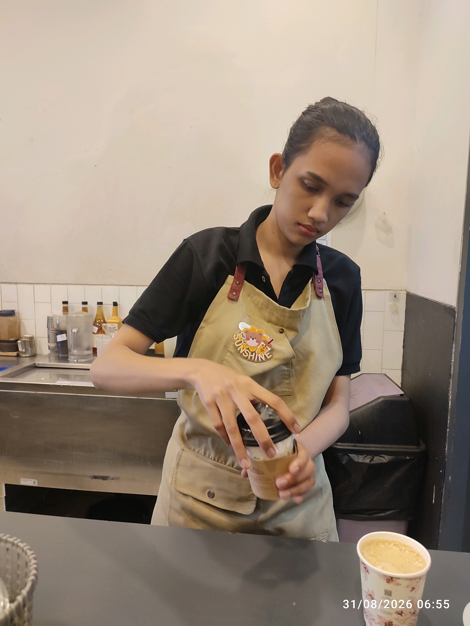 An East Jakarta, Indonesia coffee shop is depicted in this image. A young barista, wearing a black shirt and a beige apron, is in the process of preparing a coffee beverage. The apron has a cartoon bear wearing a chef's hat and the word "SUNSHINE" embroidered on it. The barista's hands are visible as they hold a paper cup, seemingly adding a final touch to the drink. In the foreground, another paper cup filled with coffee sits on a dark grey counter, accompanied by a timestamp indicating "31/08/2026 06:55". The background shows a stainless steel counter with bottles of syrup and a large plastic pitcher, as well as a black trash bin. The overall scene suggests a typical morning at a coffee establishment.