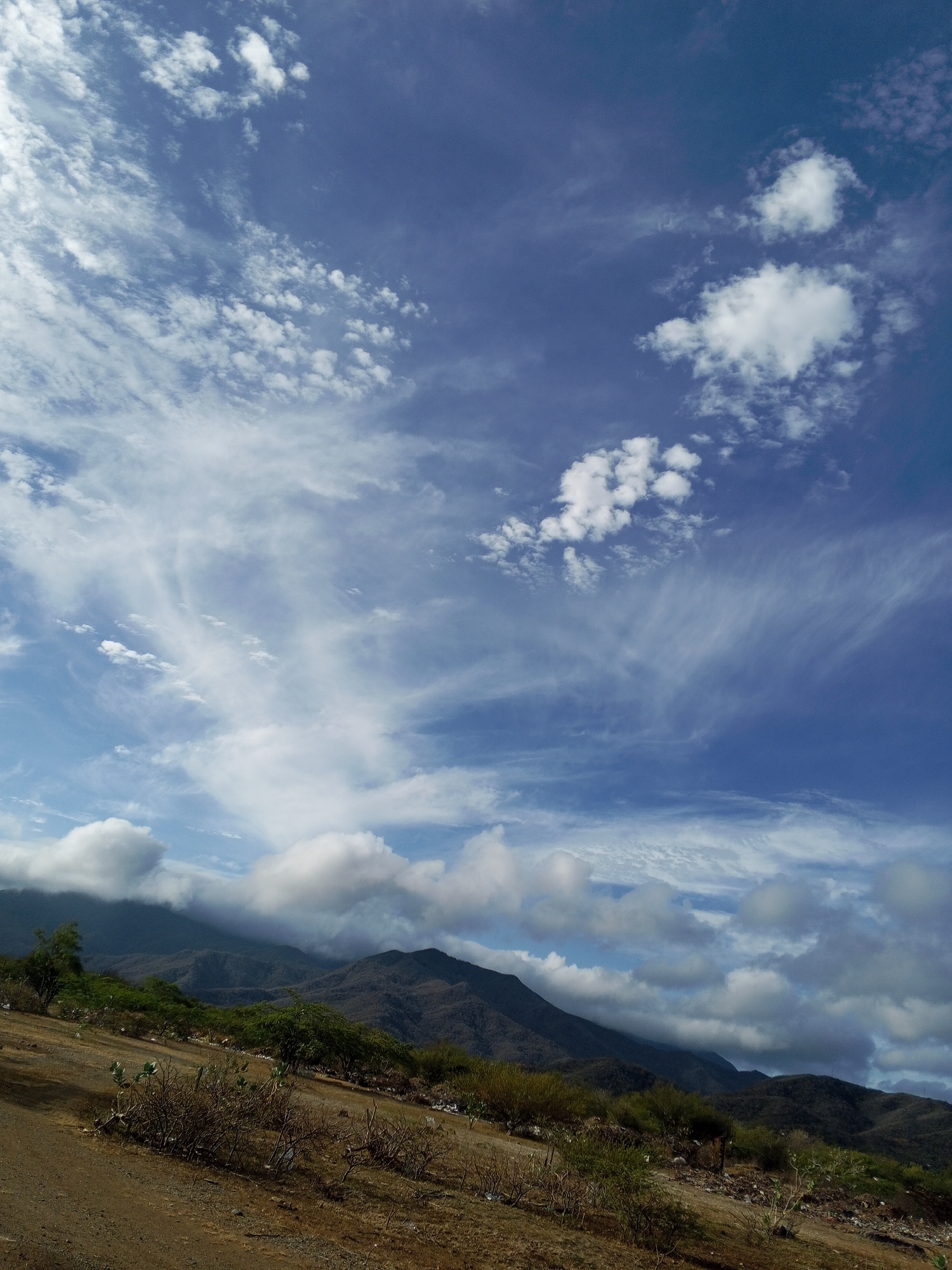 The image depicts a landscape in Altagracia, Venezuela. A mountain range is partially obscured by low-hanging clouds. The foreground shows a dirt or gravel slope with sparse vegetation. The sky is blue with a mix of cirrus and cumulus clouds. There are no people visible in the image.
