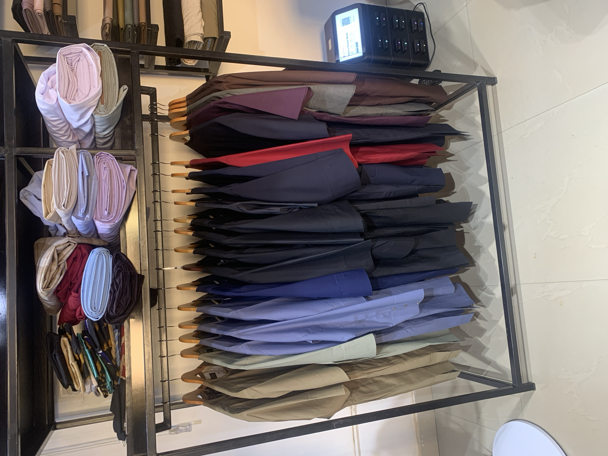 The image shows a metal rack filled with rolls of fabric and clothing items on hangers. On the left side of the rack, rolls of fabric in various colors and patterns are stacked vertically. The right side of the rack displays clothing items, potentially shirts or pants, hanging on wooden hangers, stacked one above the other. The colors of the clothing range from browns and purples at the top to blues and greens towards the bottom. At the top of the rack, a black electronic device is visible. The environment appears to be a retail or storage area, possibly a shop or warehouse. Given the request to note the location as Narayi, Nigeria, this image potentially captures the interior of a textile shop, clothing store or storage facility in Narayi, Nigeria. However, without additional details, this is speculative.